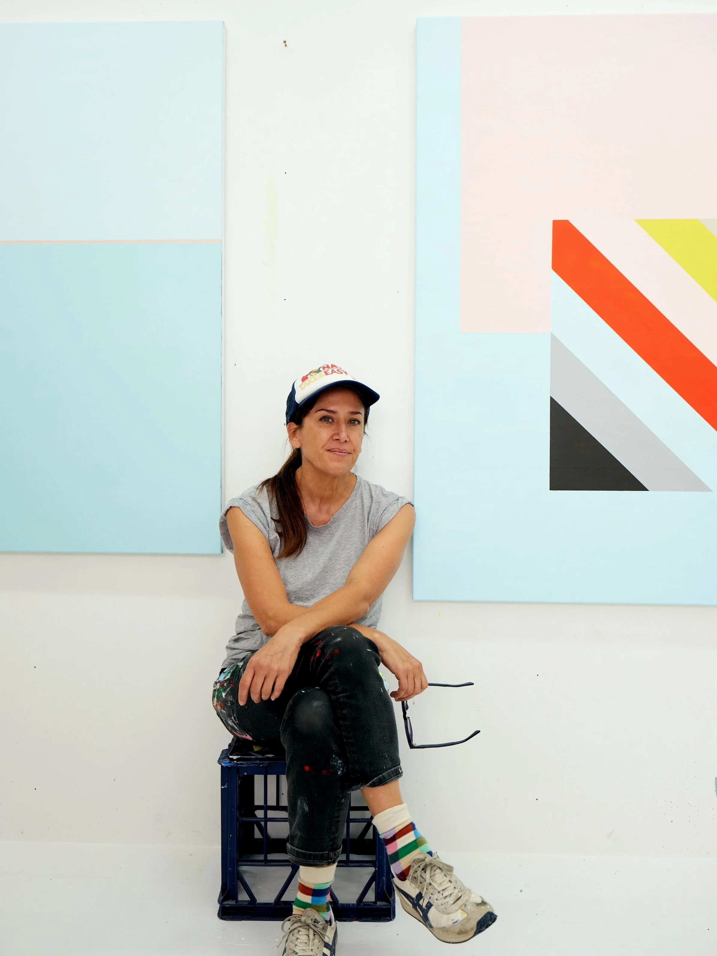 Sydney Contemporary artist Evie Adasal, sitting on a crate in her studio with minimal abstract paintings in the background.