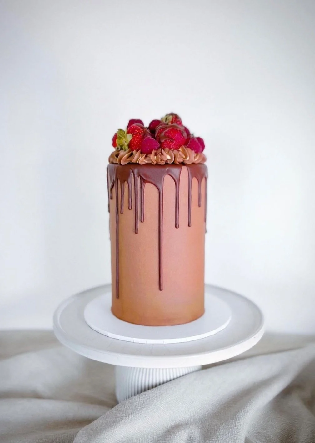 Tall layered chocolate cake with chocolate drip and topped with fresh strawberries and raspberries on a white cake stand.