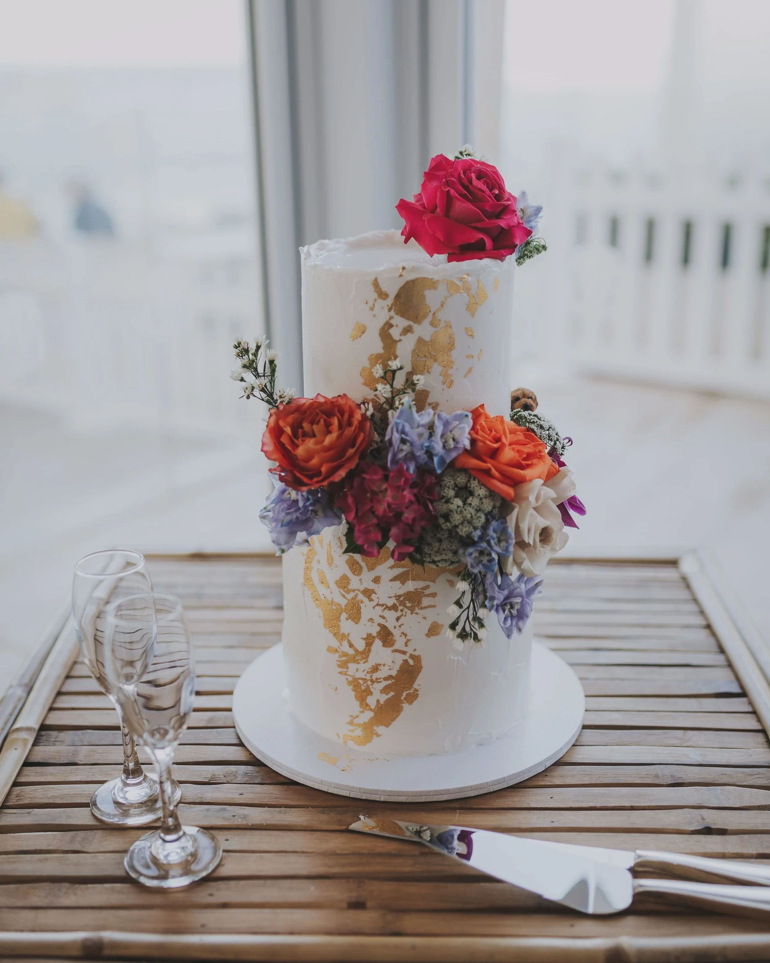 Two-tier wedding cake with white frosting, gold leaf accents, and colorful floral decorations on a wooden table. Two empty champagne flutes and a cake knife are beside it.