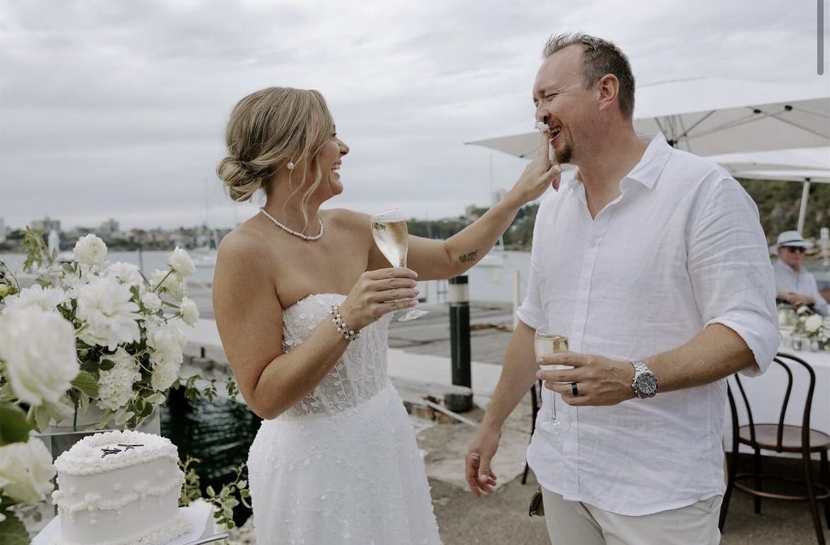 A bride and groom laughing and celebrating with glasses of champagne beside a decorated cake at an outdoor wedding reception, with a waterfront view.