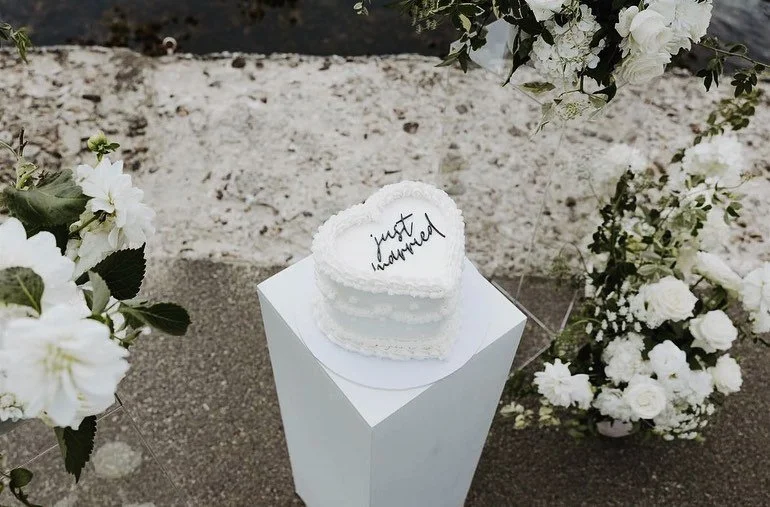 Heart-shaped "just married" cake on a white pedestal surrounded by white flowers outdoors.