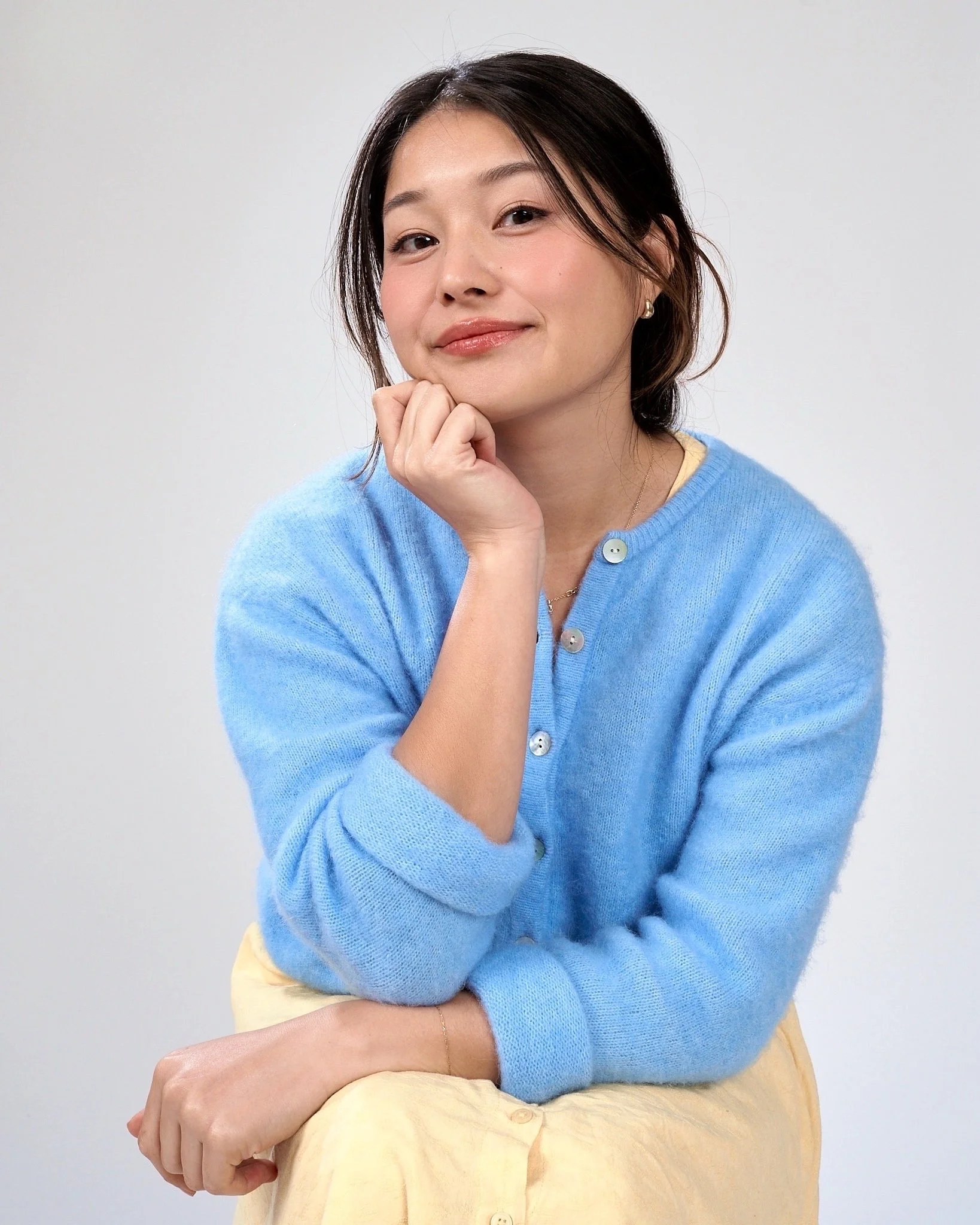 Korean American female therapist wearing a blue cardigan and yellow dres smiles at the camera with a hand on her chin.