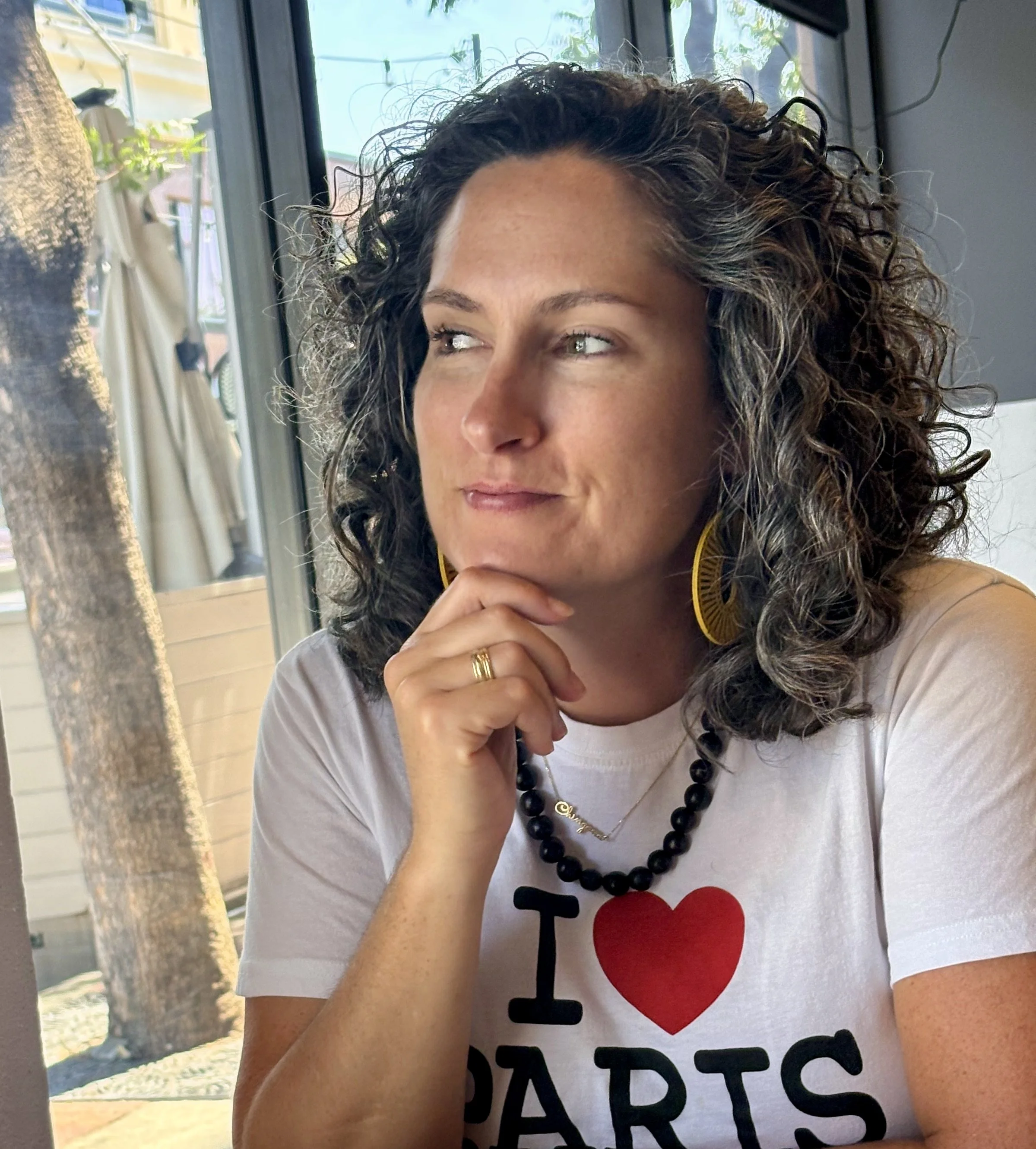 A woman with curly dark hair and light skin sitting at a table, looking thoughtfully out of the window. She is wearing a white t-shirt that says 'I ♥ PARTS,' large yellow earrings, a black beaded necklace, and a ring on her finger.