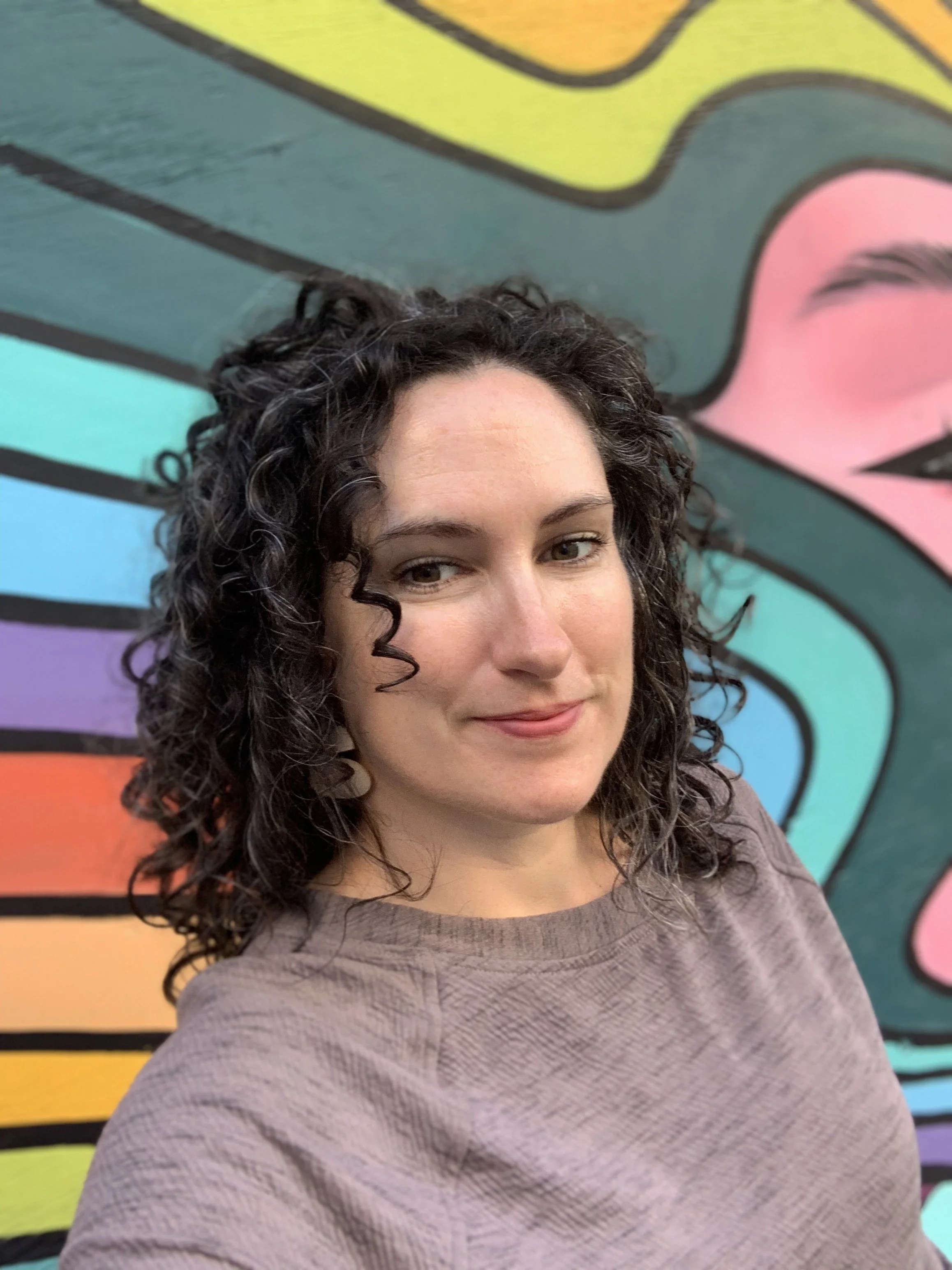 A woman with curly dark hair and earrings taking a selfie in front of a vibrant, colorful mural.