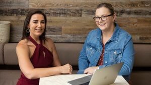 Two women sitting at an outdoor cafe table with a laptop, smiling, professional headshot by Cole Studios.
