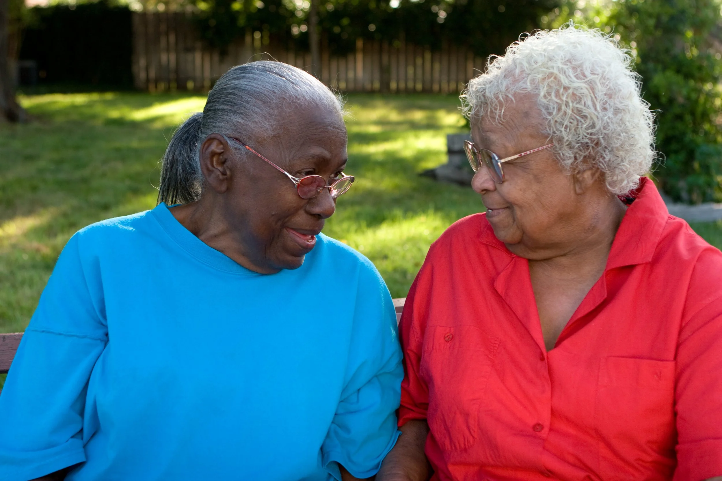 Two elderly women with glasses smiling and talking to each other, sitting outdoors on a bench in a park with green trees and grass.
