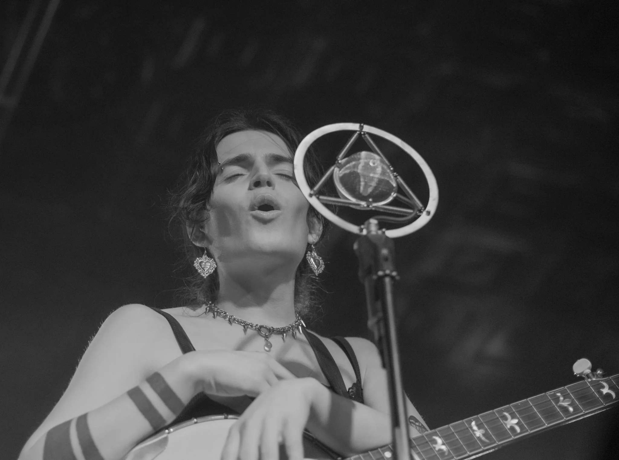 A woman singing into a vintage microphone while playing guitar, dressed in a dark sleeveless top and jewelry, with short dark hair and expressive pose.