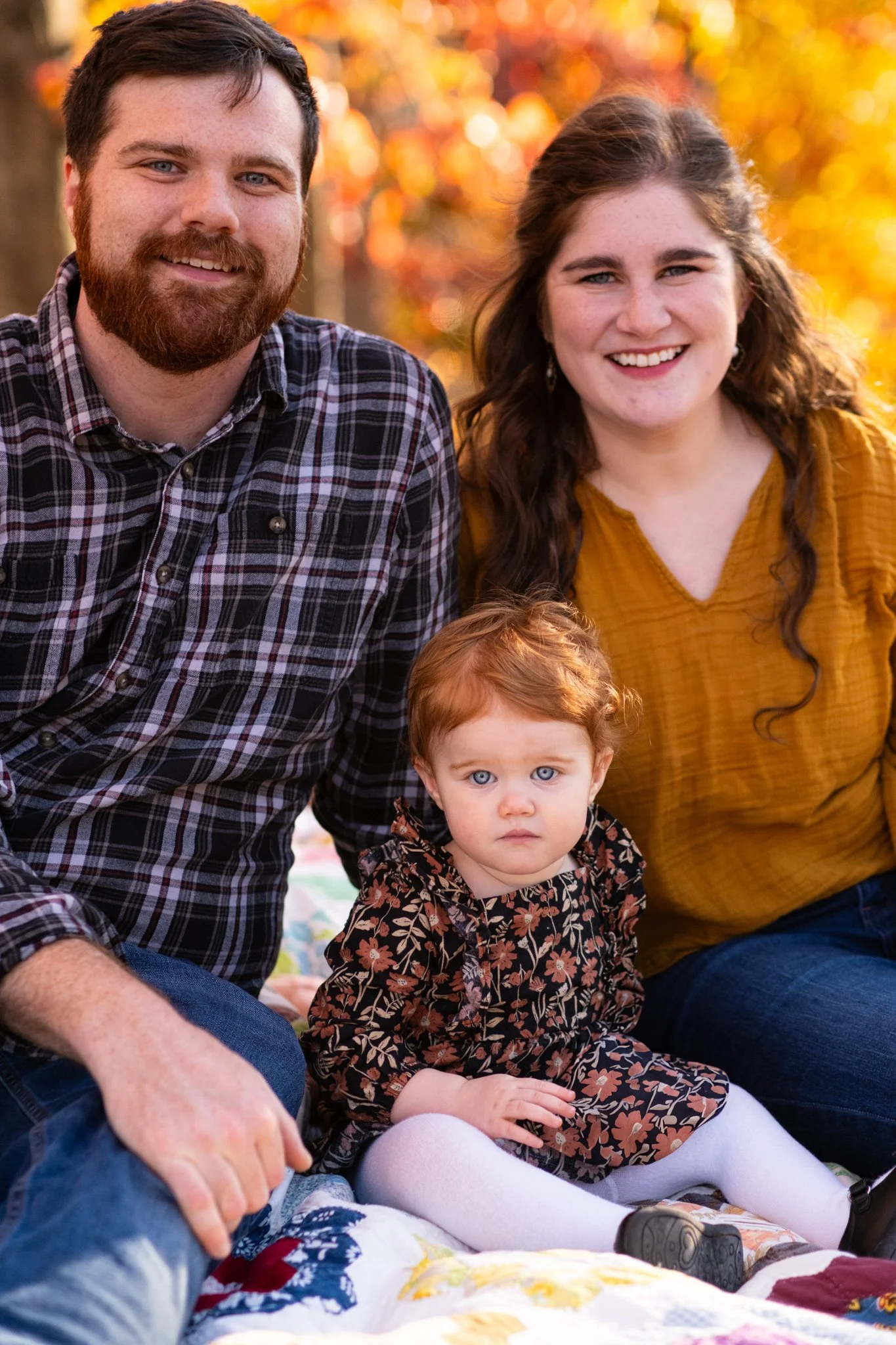 Family of three sitting on a quilt outdoors during fall, with colorful autumn leaves in the background. The man has a beard and is smiling. The woman has long wavy hair and is smiling. The young girl has curly red hair and is looking serious.