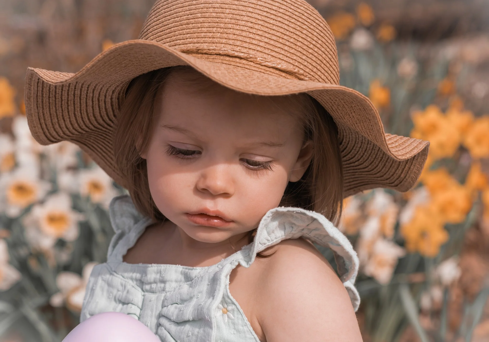 A young girl wearing a large straw hat and a light-colored sleeveless top looks down with a thoughtful expression in a garden of yellow and white flowers.