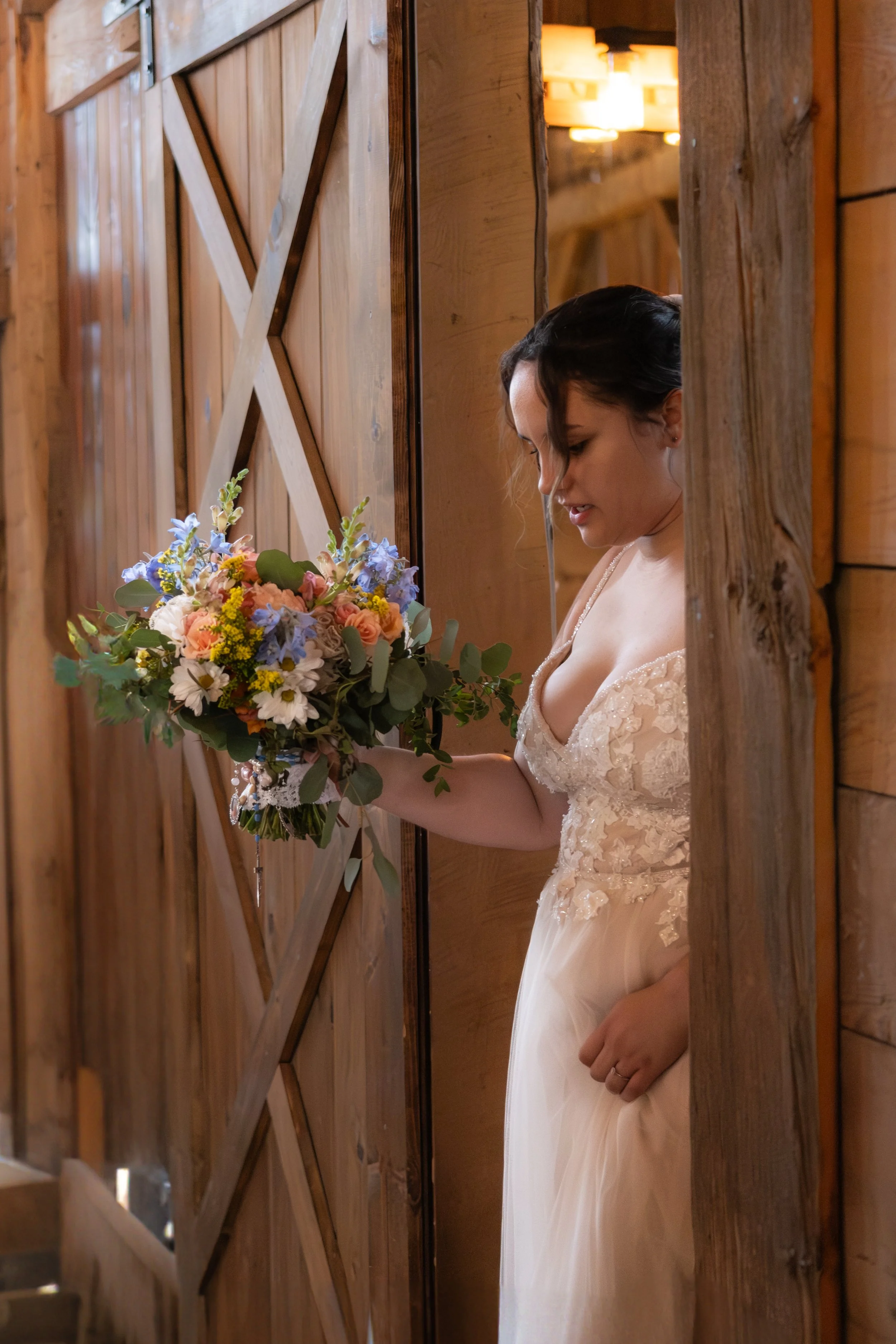 A woman in a wedding dress holding a bouquet of flowers, standing beside a wooden barn door.