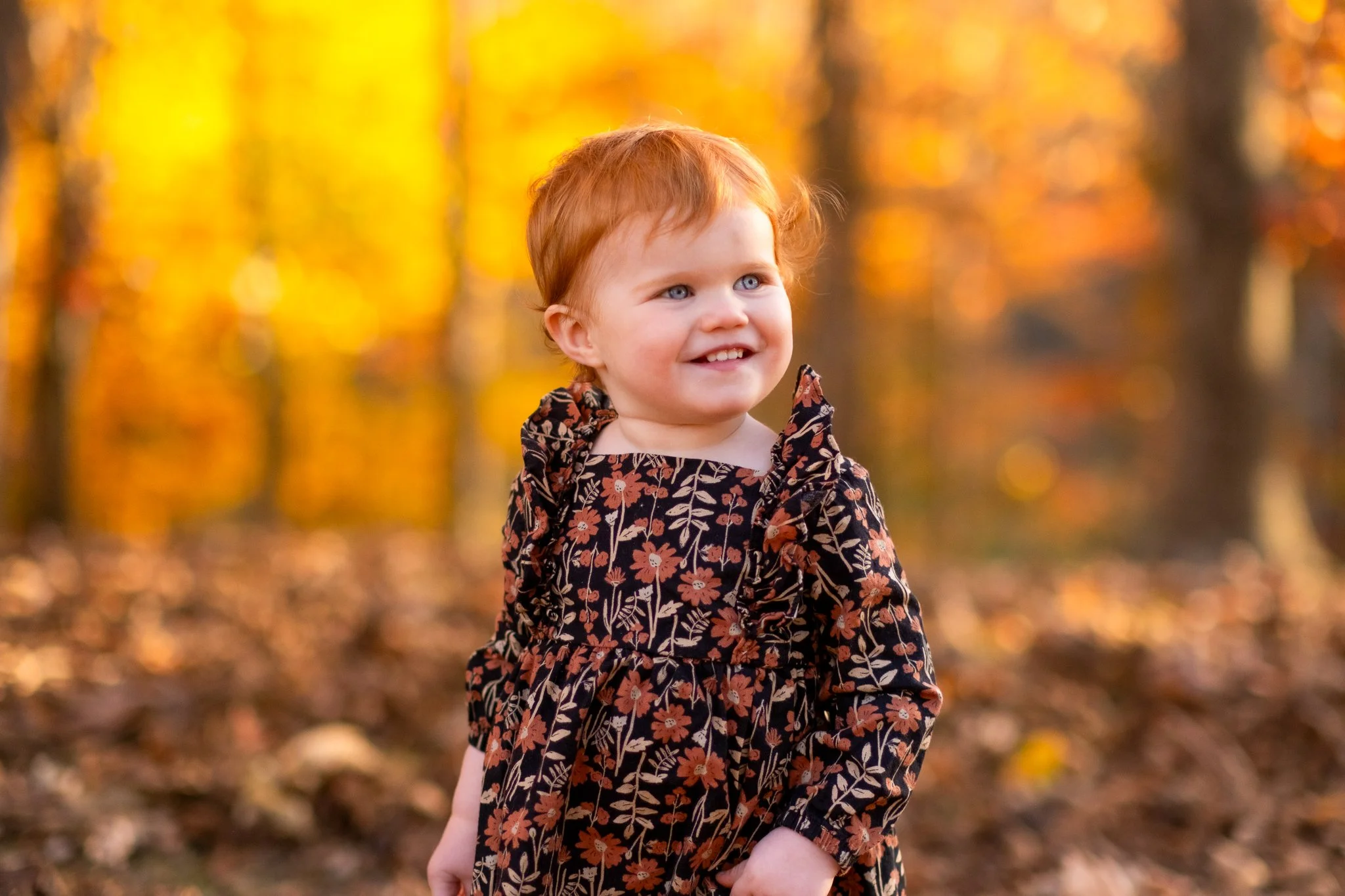 A young girl with red hair and blue eyes smiling outdoors in a fall setting with orange and yellow leaves in the background.