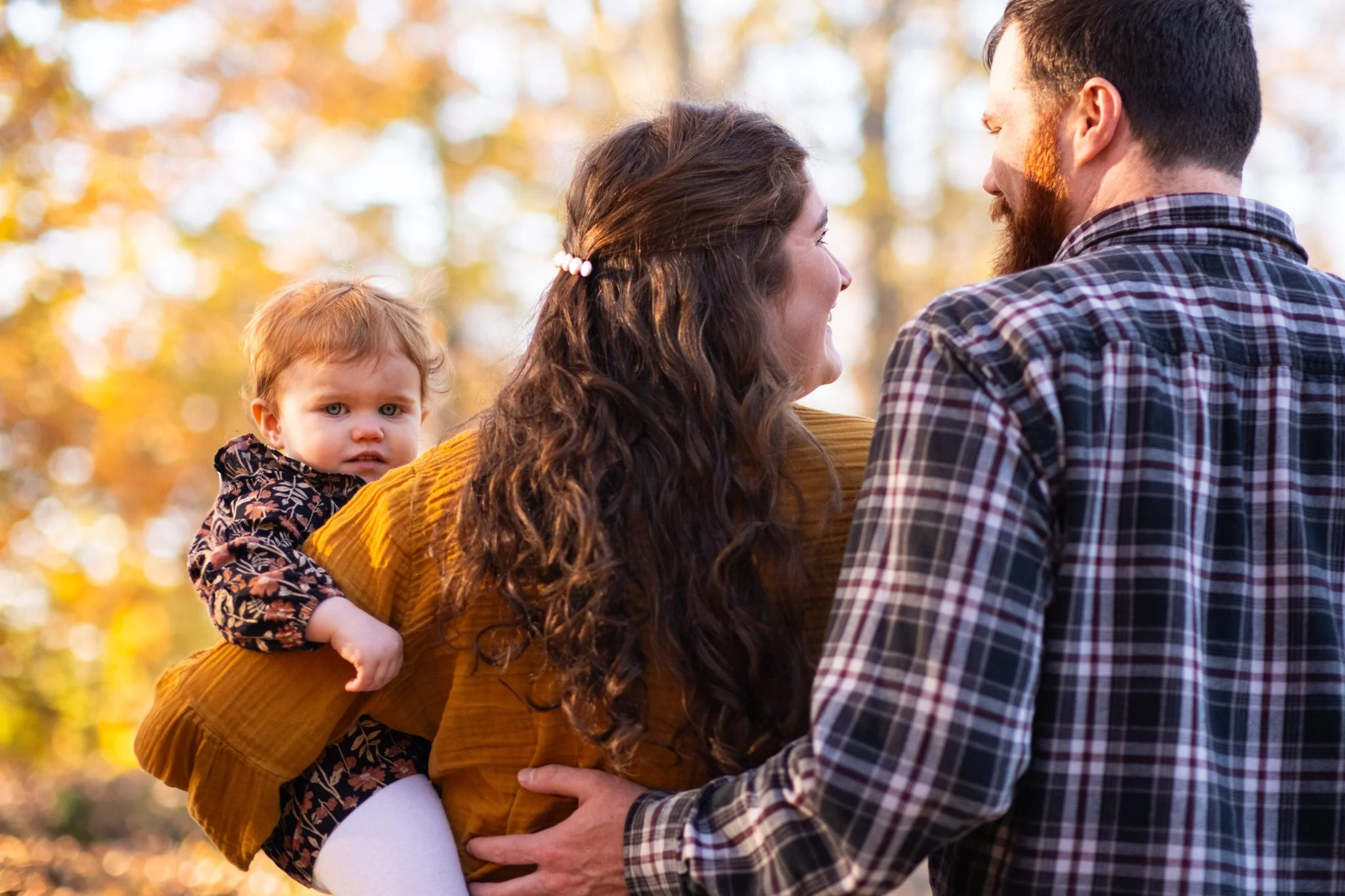 A family of three outdoors in autumn, with a woman with long curly brown hair smiling at a man with a beard and short dark hair, while a toddler with curly blonde hair and blue eyes looks at the camera from her back, all surrounded by fall-colored tr