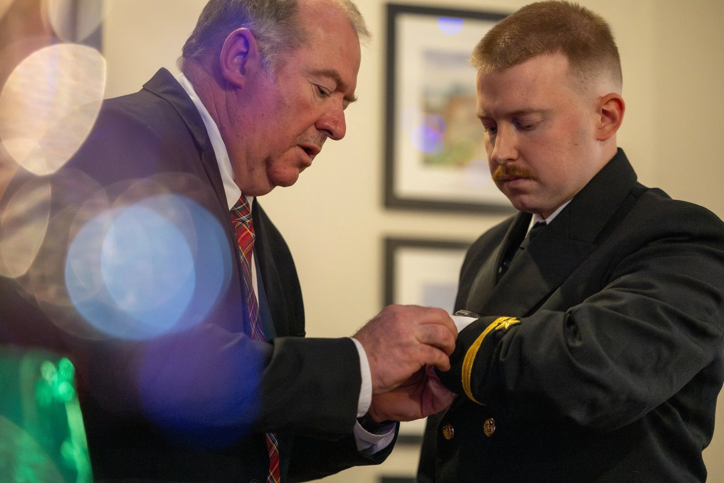 An older man in a suit and tie adjusting the cuff of a younger man in a police uniform inside a building.