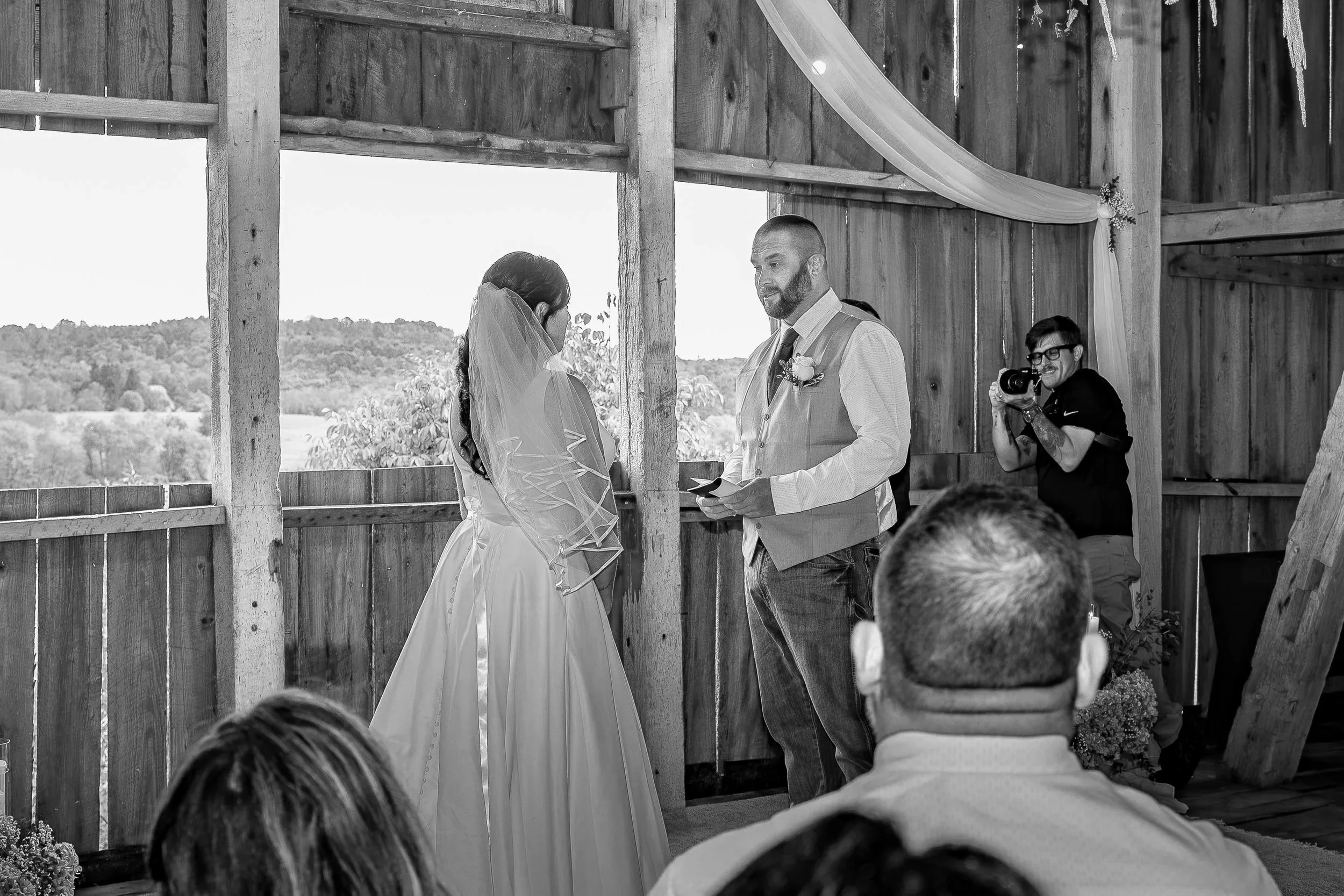 A black and white photo of a couple exchanging vows during a wedding ceremony inside a rustic wooden barn, with a photographer capturing the moment.