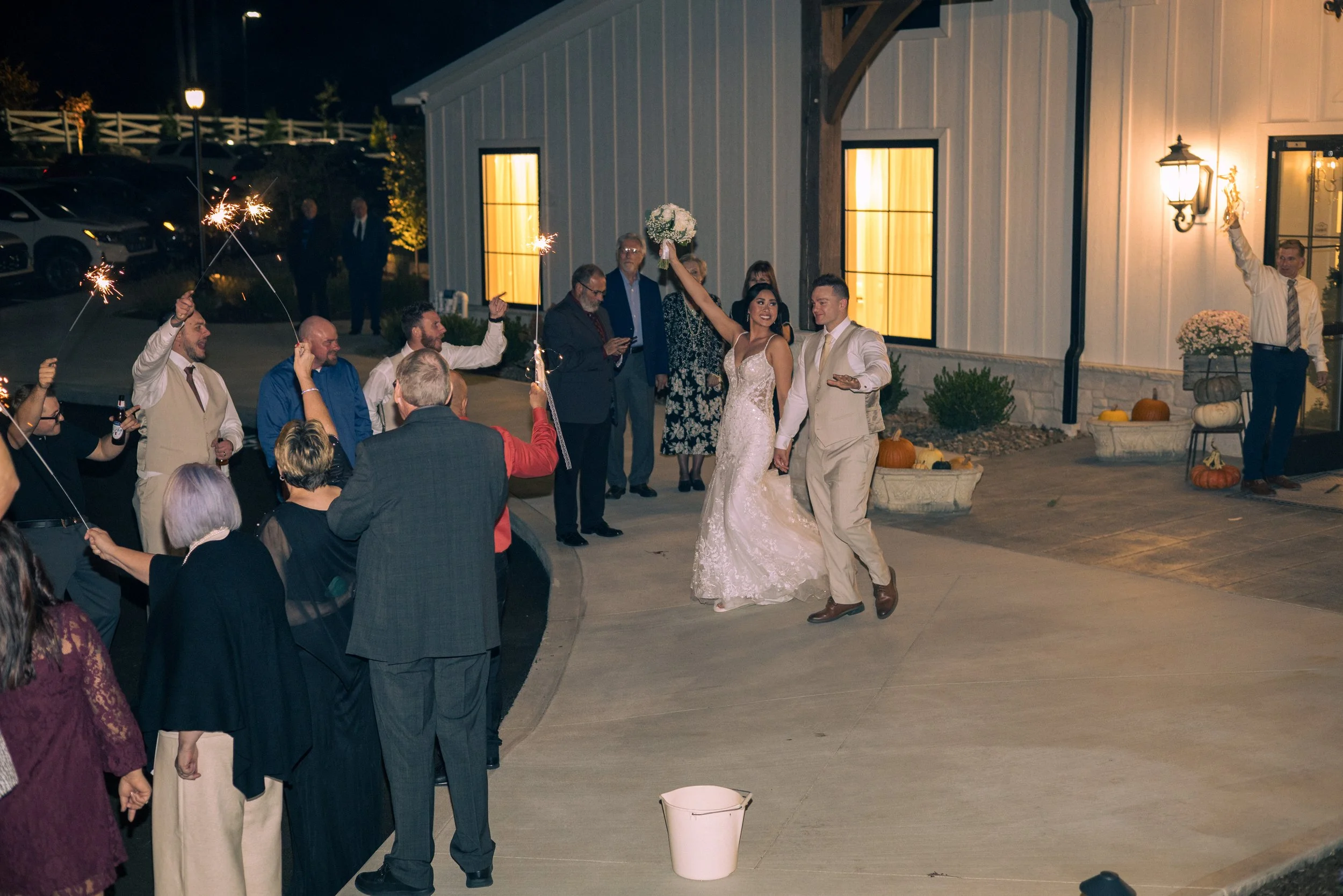 A newlywed couple dancing outside at night, surrounded by guests holding sparklers. The bride is holding a bouquet and is smiling, while the groom is in a light-colored suit. There are pumpkins and fall decorations by a white building with illuminate