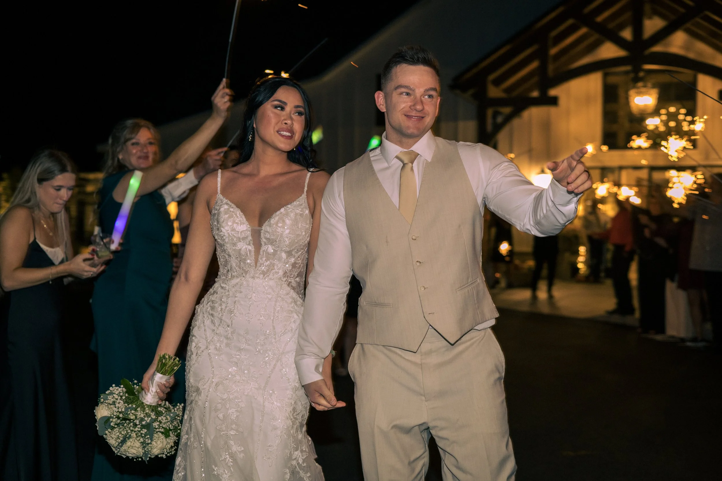 A newlywed couple celebrating their wedding with guests and sparklers at night, outdoors.