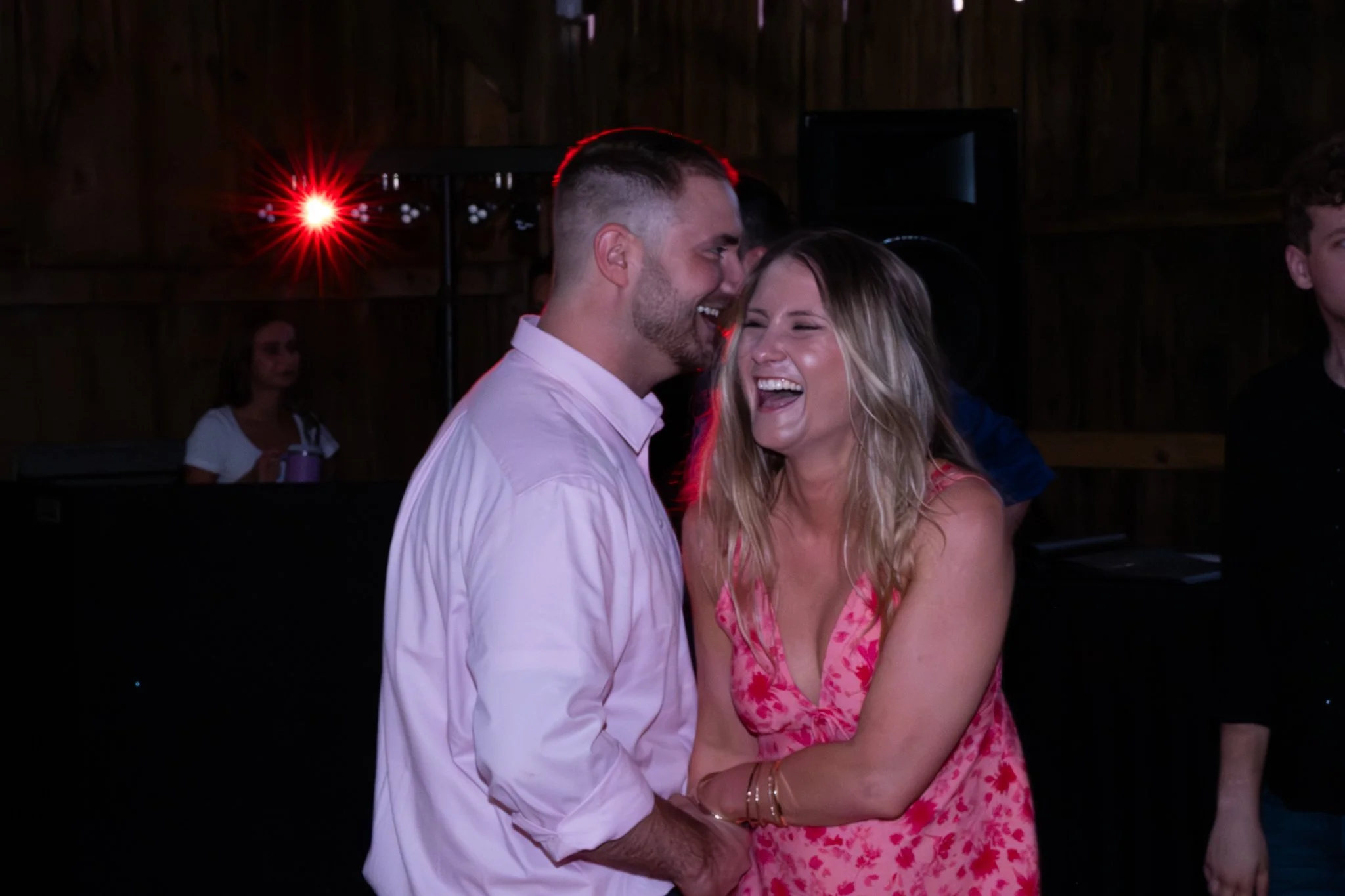 A man and a woman are dancing together in a dimly lit room, smiling and laughing, with a red light shining in the background.