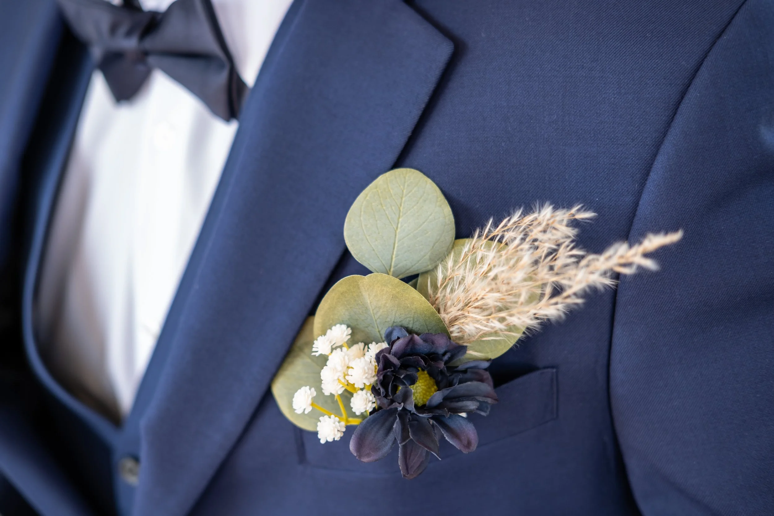 A boutonniere with white, black, and yellow flowers, dried grass, and green leaves pinned to a dark navy suit jacket, with a white shirt underneath.