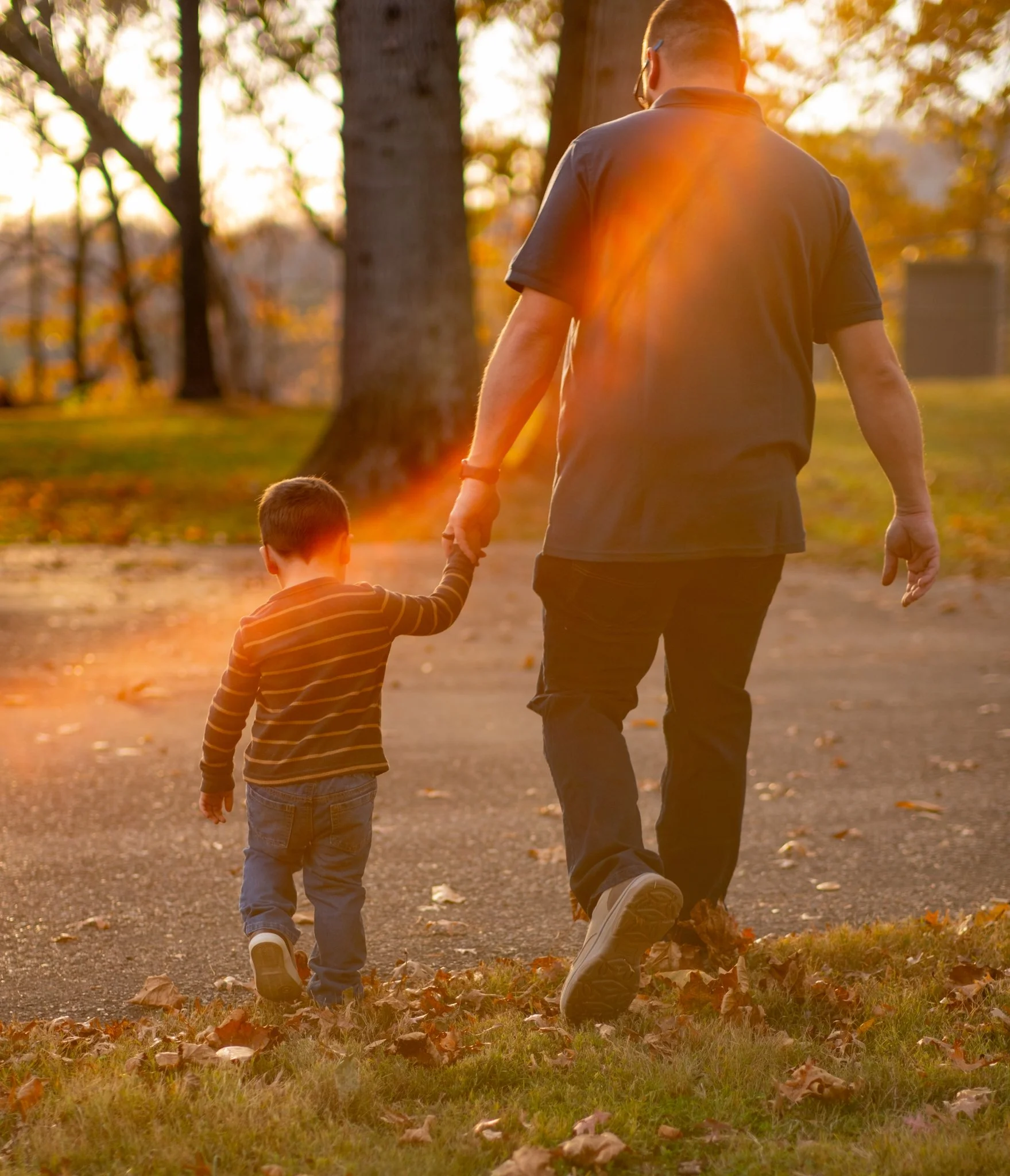 An adult and a young boy walking hand in hand outdoors during autumn, with trees and fallen leaves on the ground, captured at sunset.