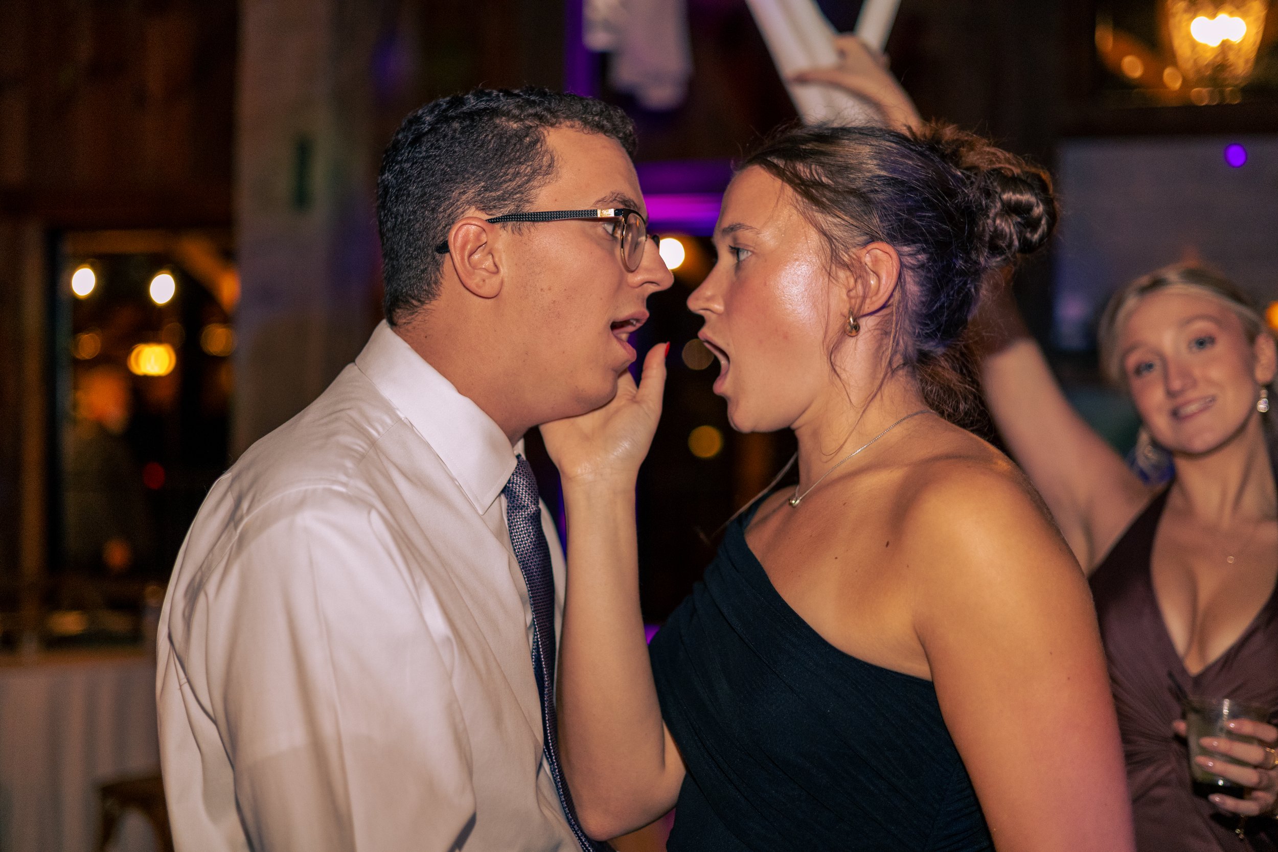 A man with glasses and a woman in a strapless dress are close, touching foreheads with mouths open, in a dimly lit party or celebration setting.