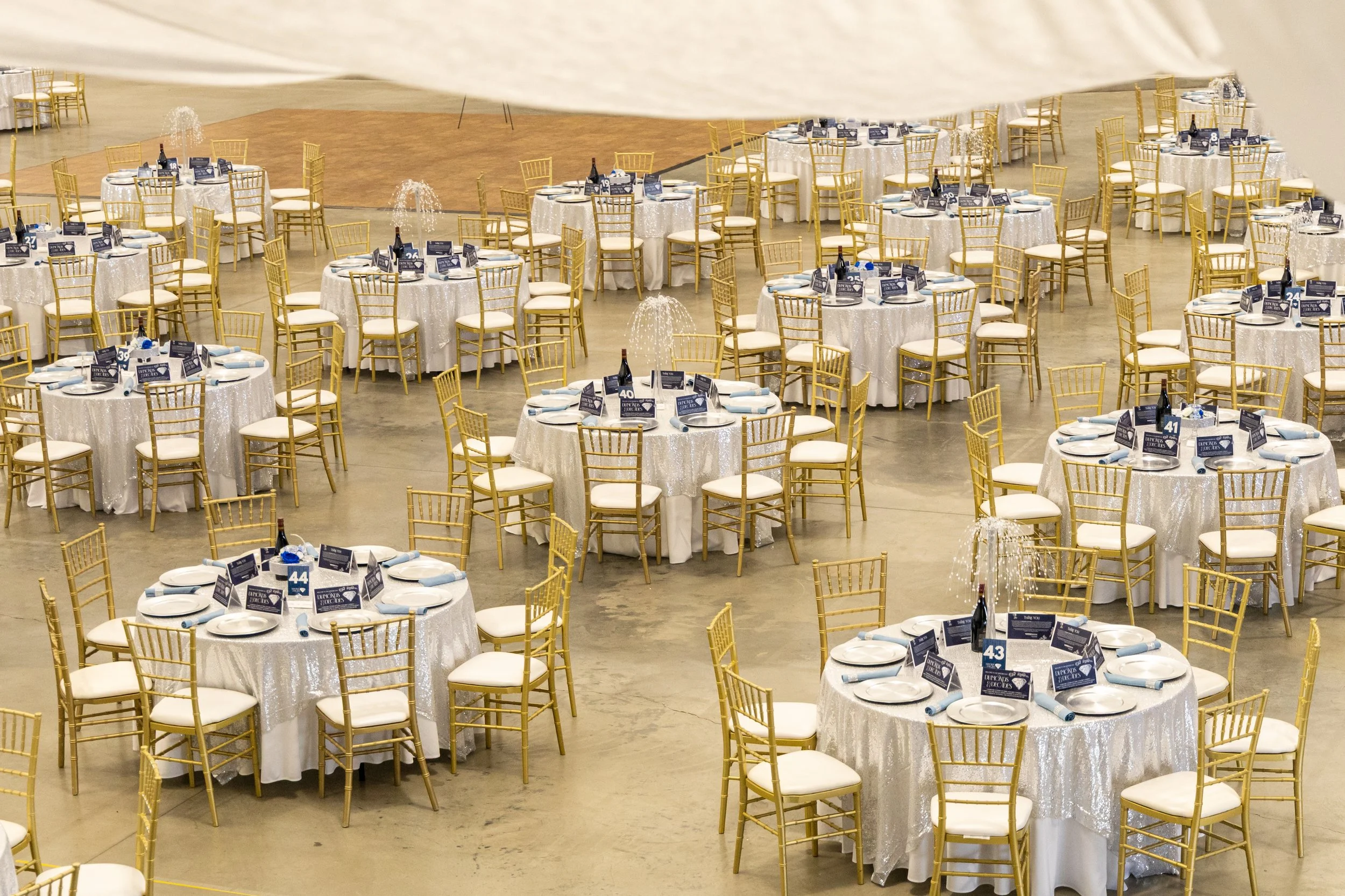Multiple round banquet tables with white tablecloths and gold chairs within a large event hall, set for a formal event.