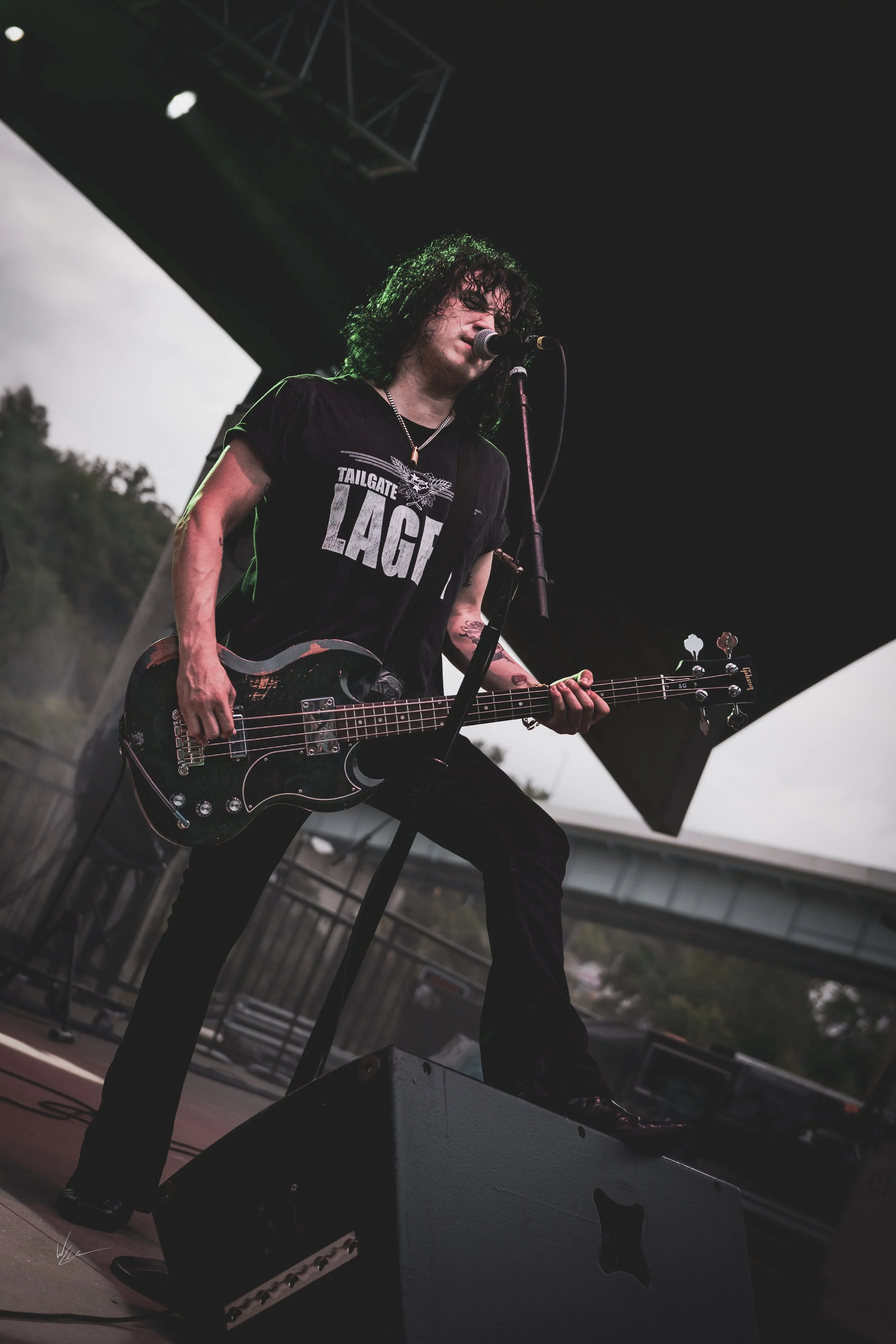 Musician with black curly hair playing bass guitar and singing into a microphone on an outdoor stage.