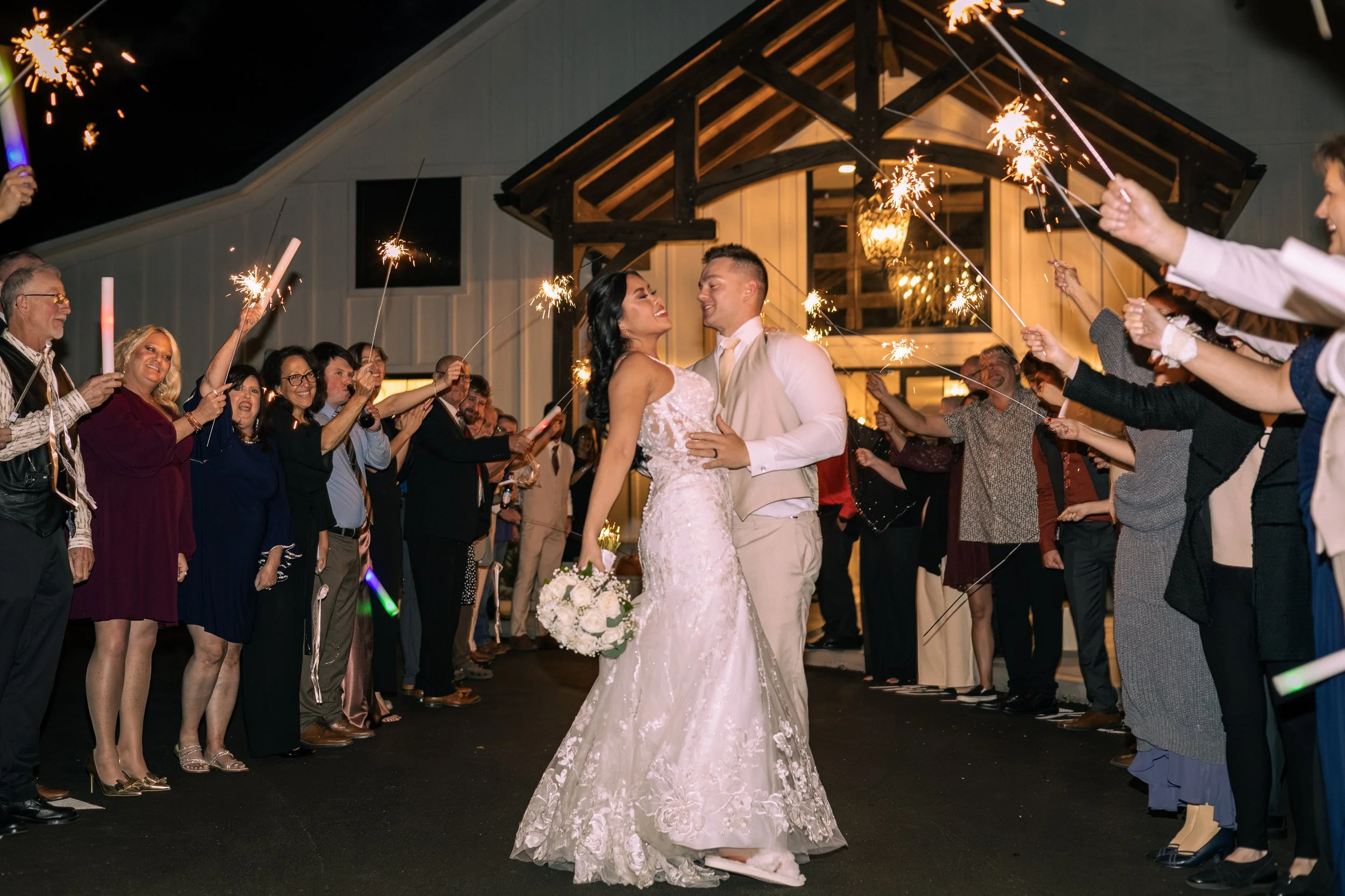 Bride and groom dancing at their wedding reception surrounded by family and friends holding sparklers at night outside a barn-style building.