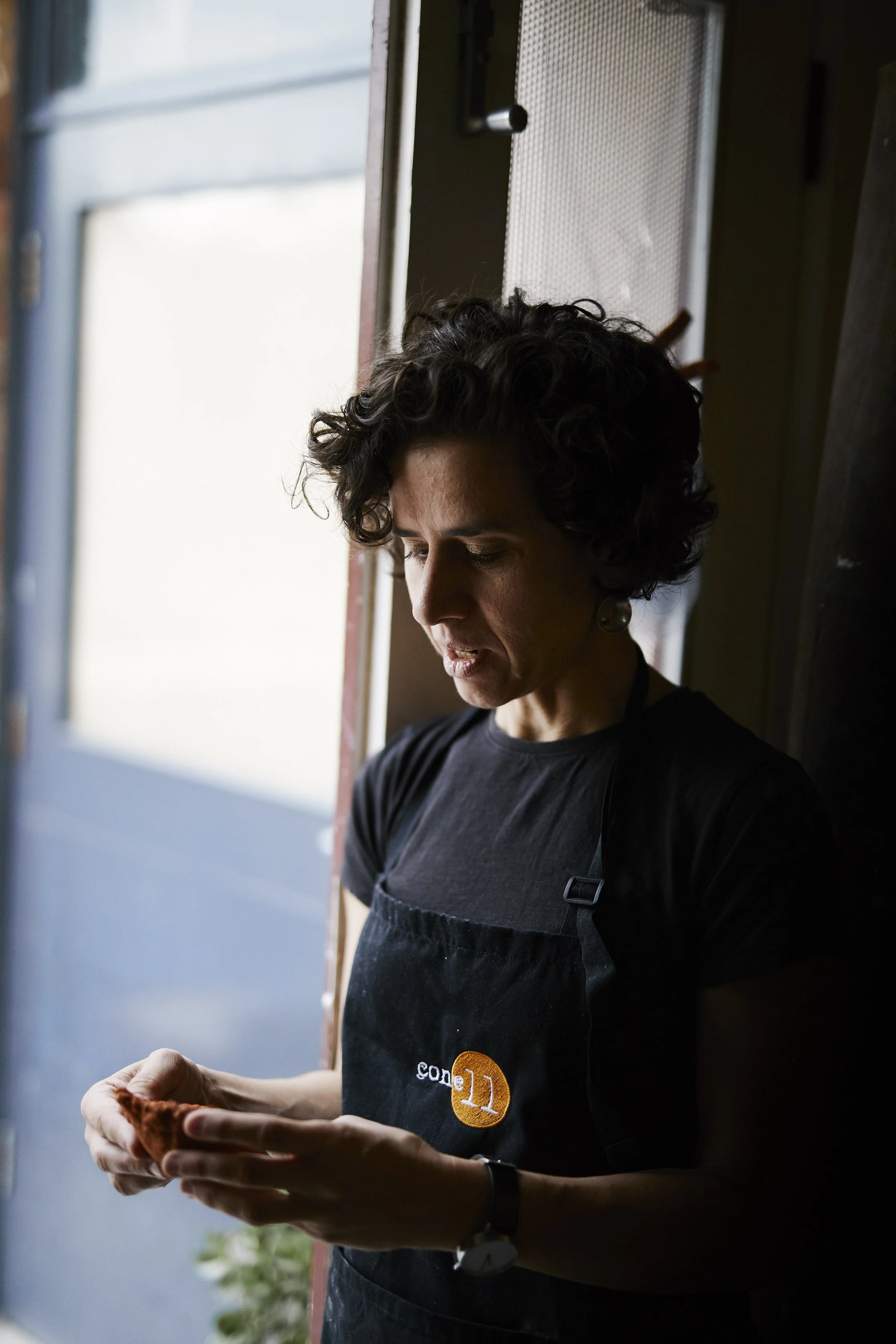A woman with short, curly dark hair looking down at her hands, wearing a black t-shirt and a black apron with a small round logo, standing near a doorway with natural light coming in.
