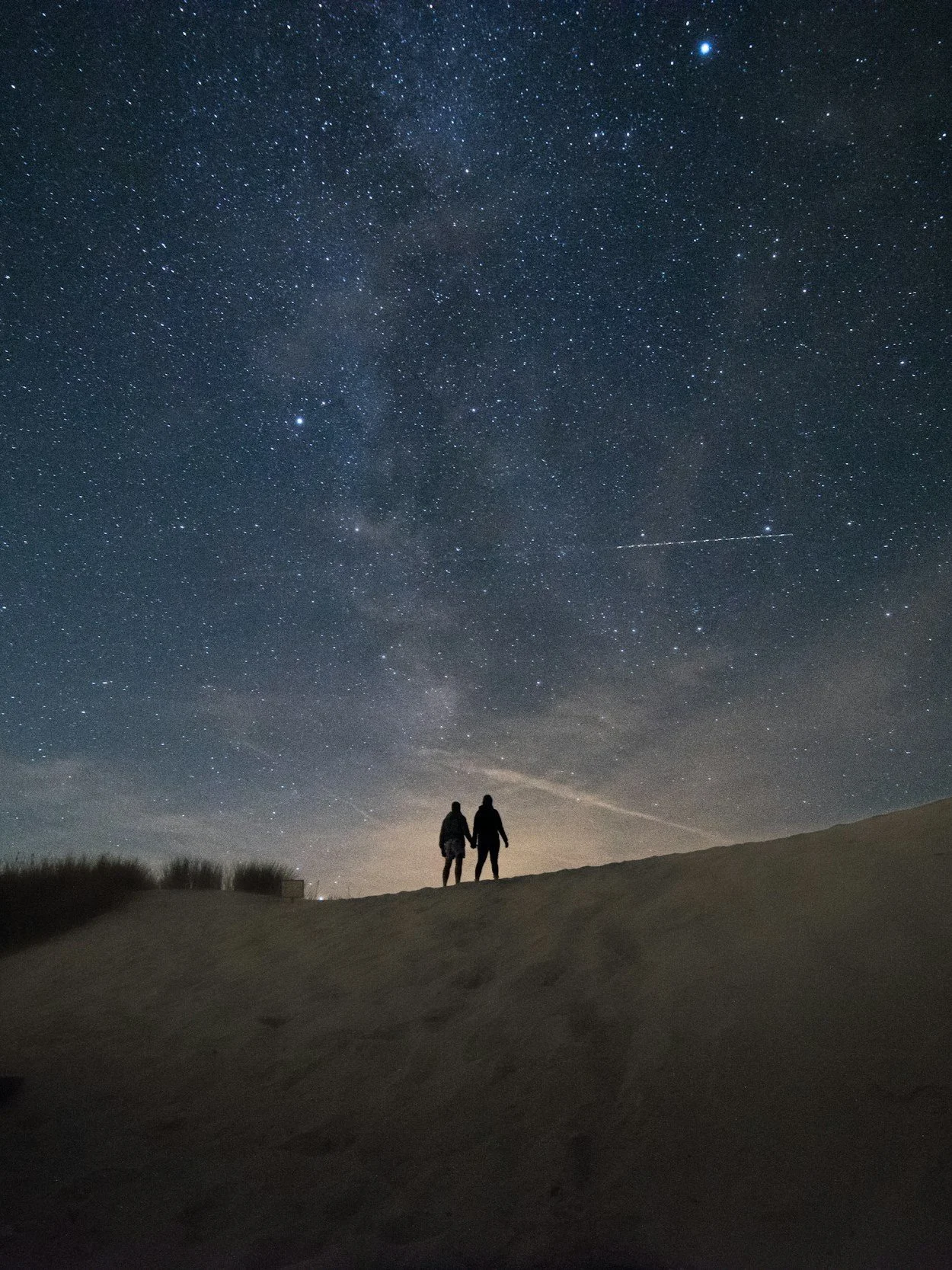 Silhouettes of two people holding hands standing on a hilltop under a starry night sky with the Milky Way galaxy visible.
