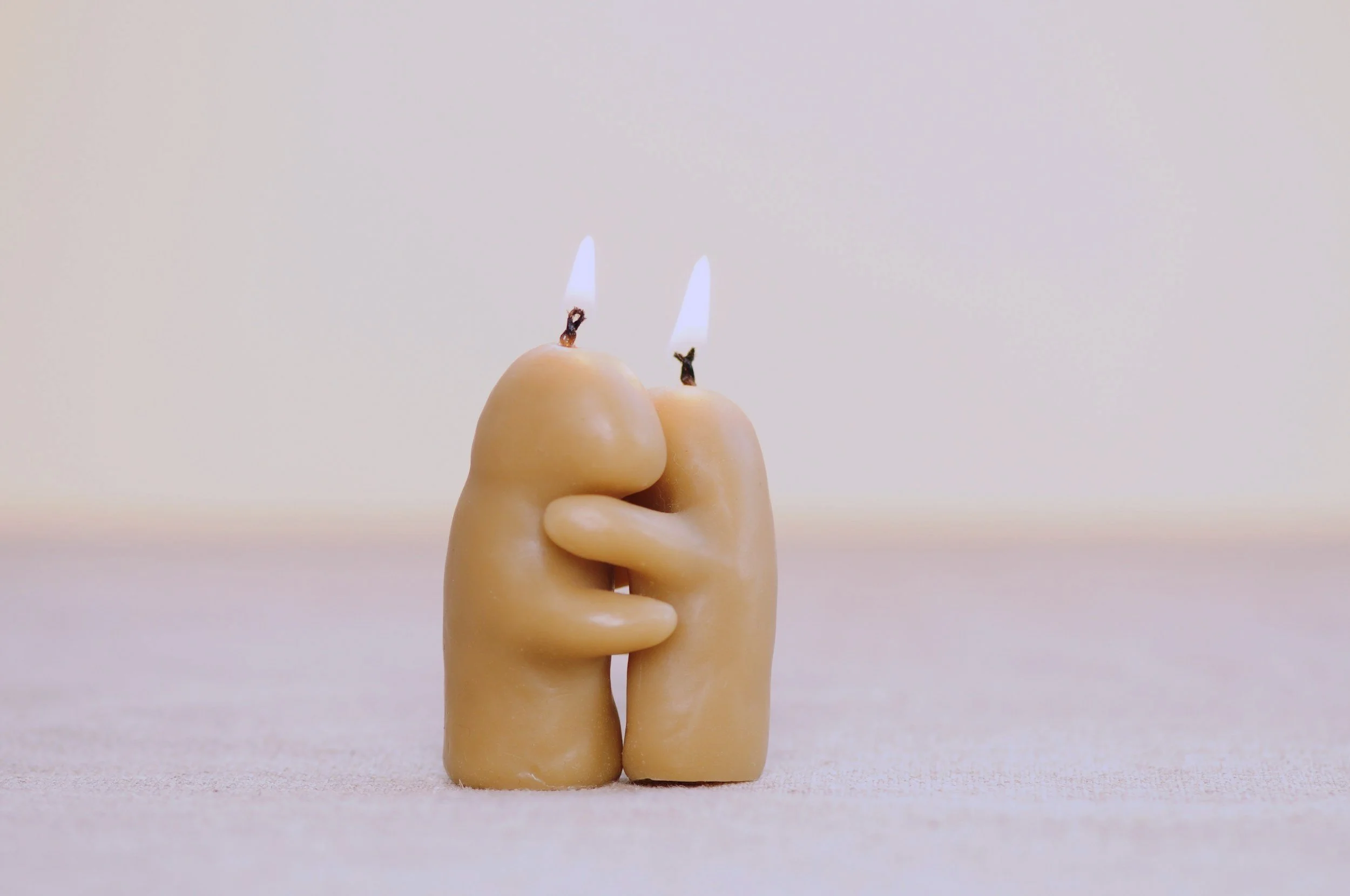 A beige candle shaped like two little figurines holding each other in an embrace, with flames on top of their heads, on a neutral surface.