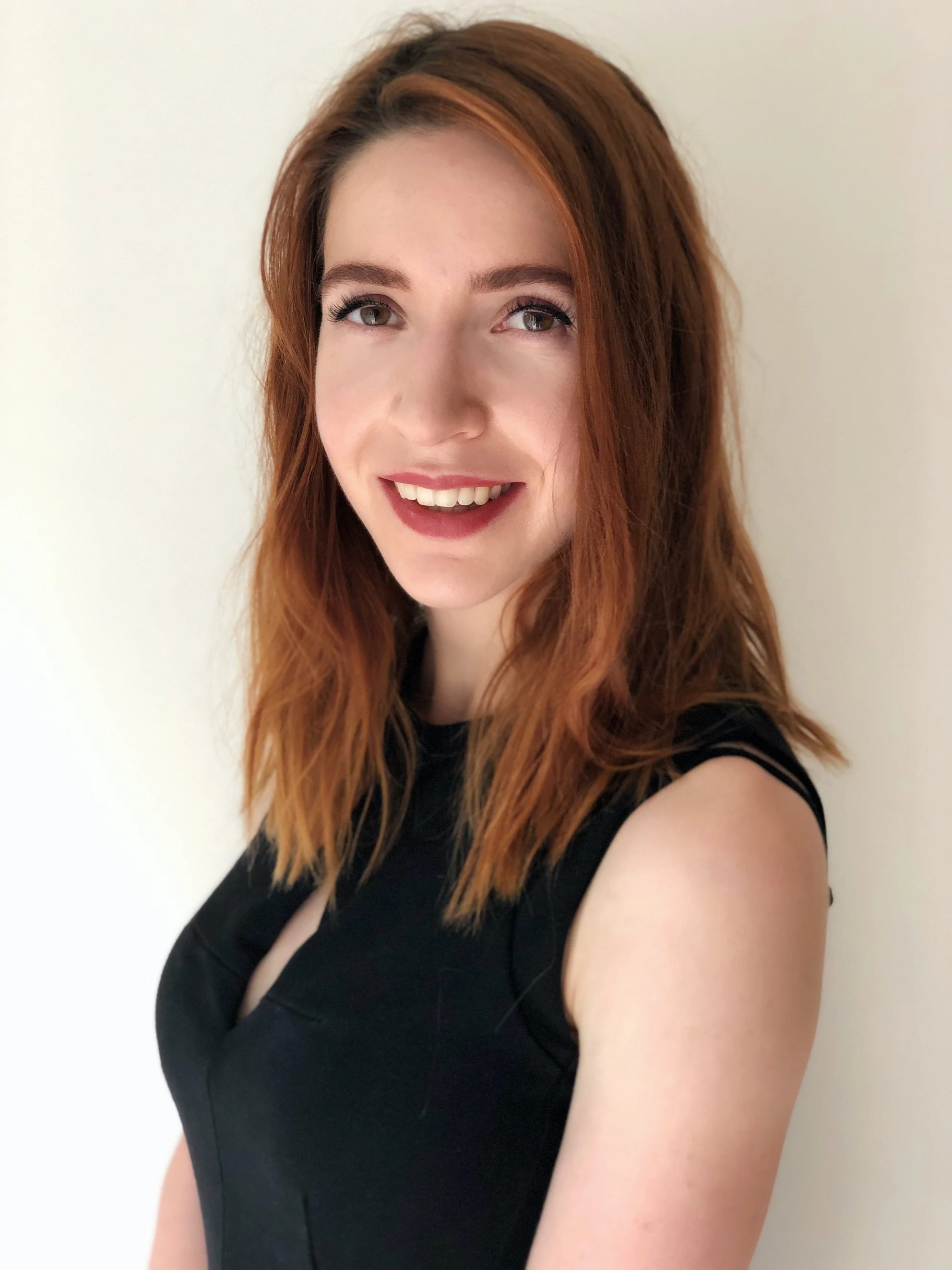 A young woman with shoulder-length reddish hair, wearing a black sleeveless top, smiling, standing against a plain off-white background.