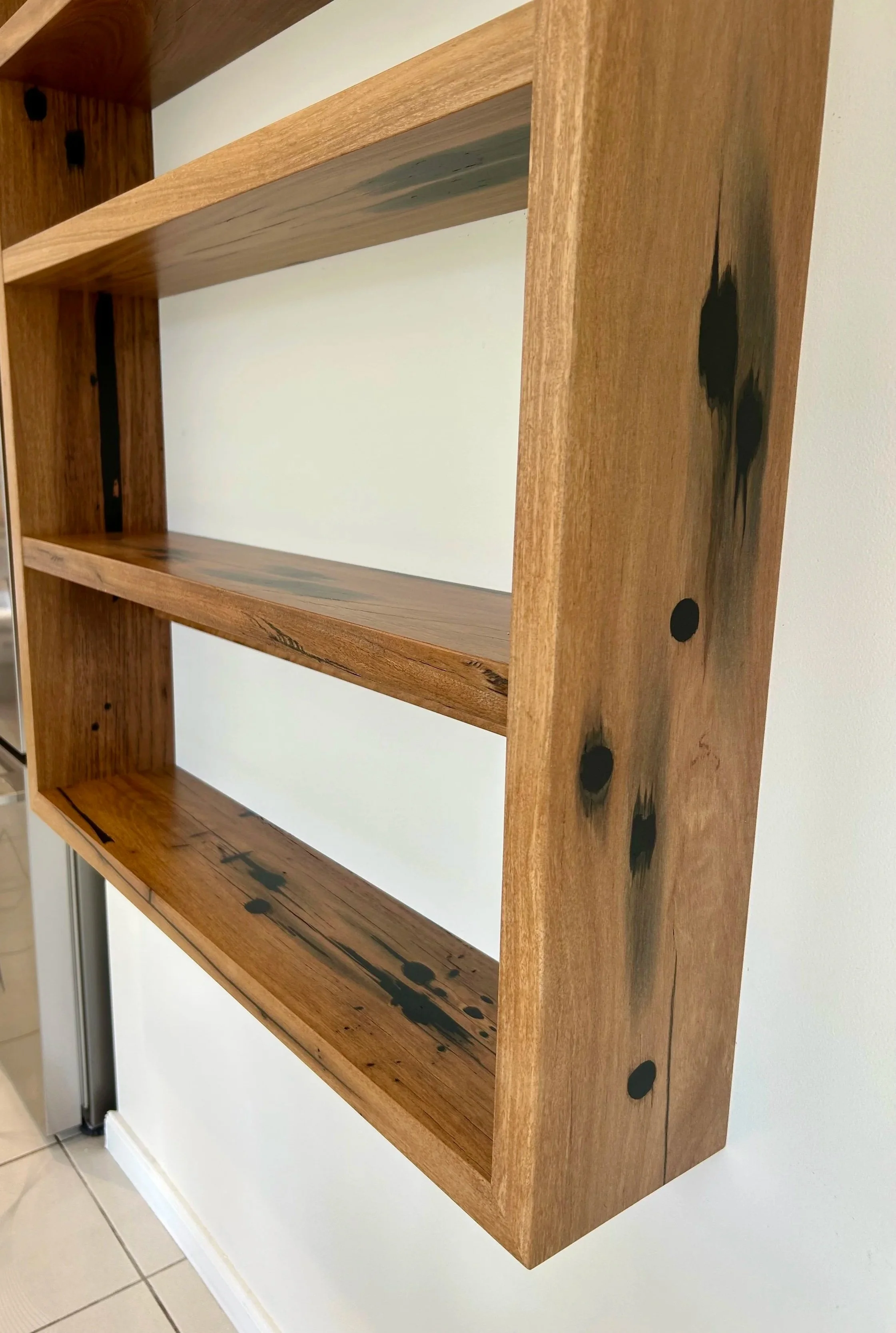 Close-up of a wooden corner shelf with black paint splatters on the side, mounted on a white wall. The shelf has a natural wood finish with visible grain and knots.