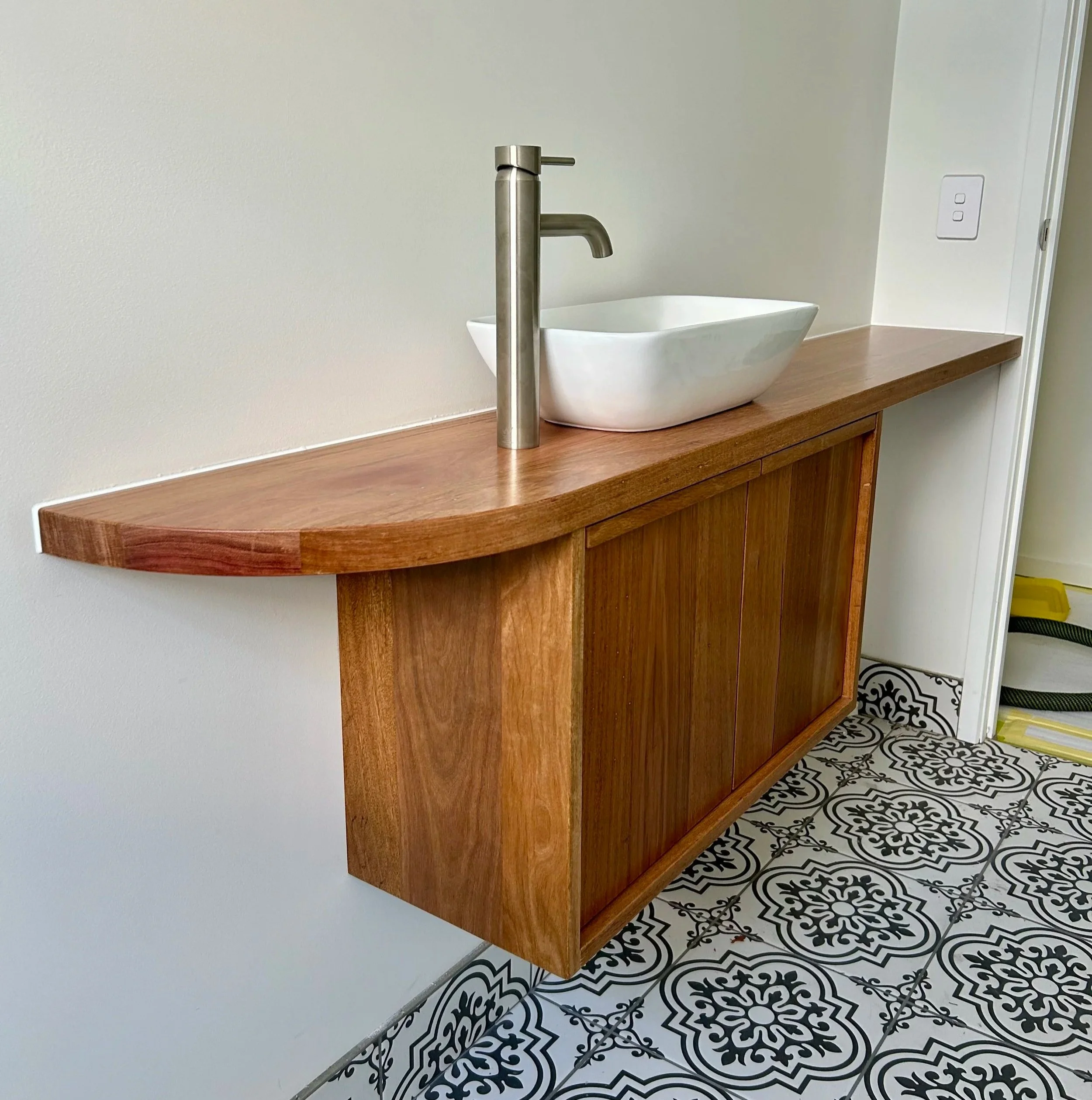 A modern wall-mounted wooden vanity with a rounded front edge, a white rectangular vessel sink, and a brushed nickel faucet, set against a white wall with patterned black and white floor tiles.