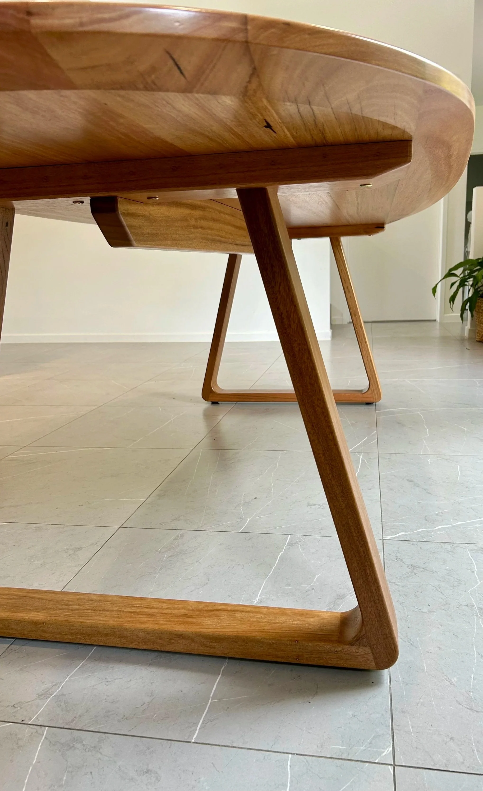 Close-up view of wooden table legs and part of the tabletop, showing a smooth finish and rounded edges, with a tiled floor and a potted plant in the background.