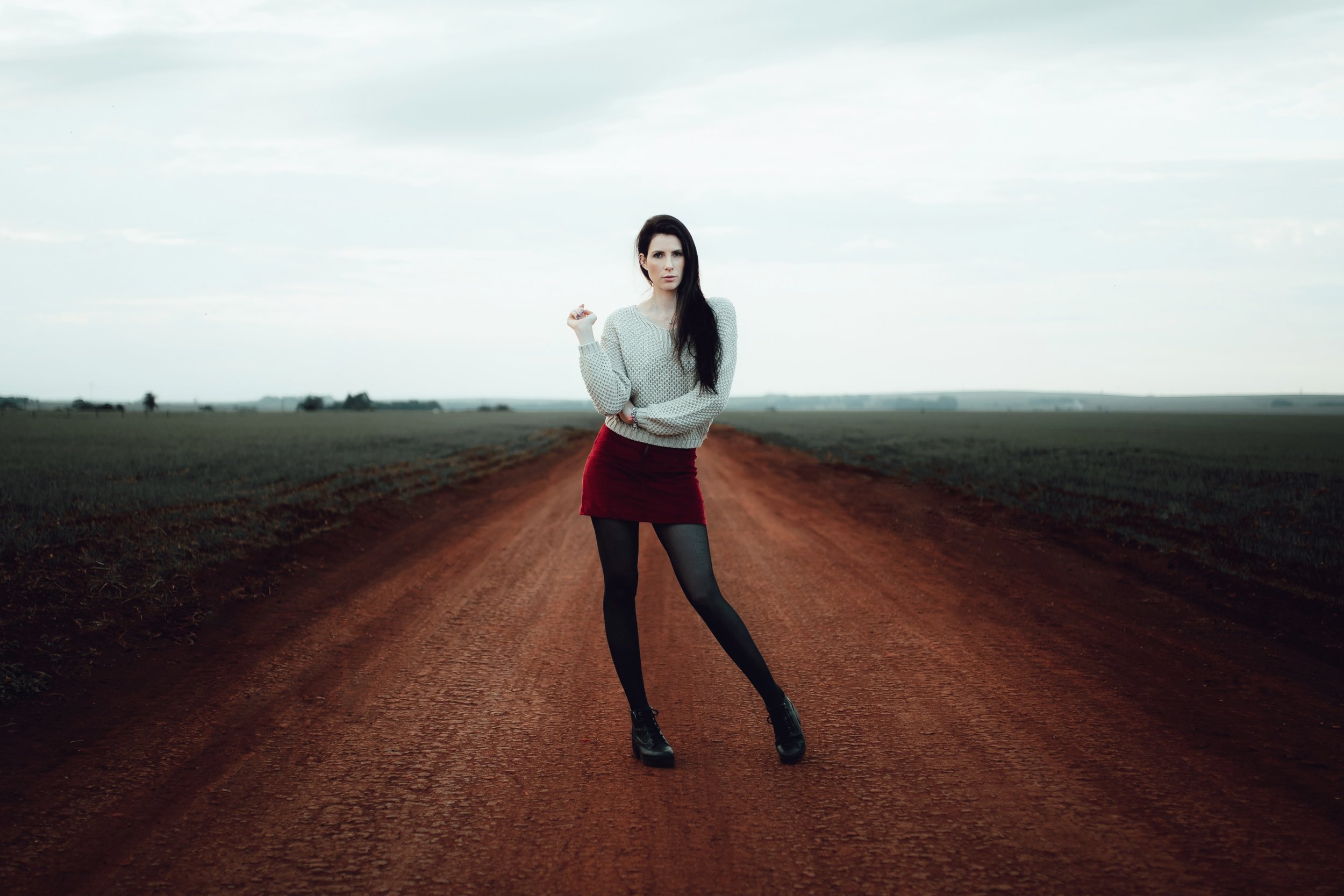 Mulher em pé em uma estrada de terra cercada por campos, usando suéter cinza, saia vermelha e meias pretas.
