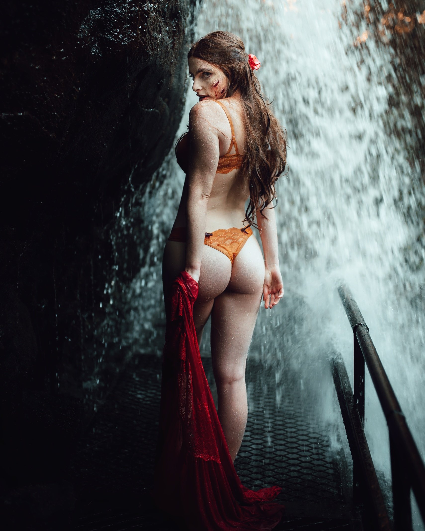 Mulher de costas, com cabelo longo, usando lingerie laranja em frente a uma cachoeira, segurando um tecido vermelho.