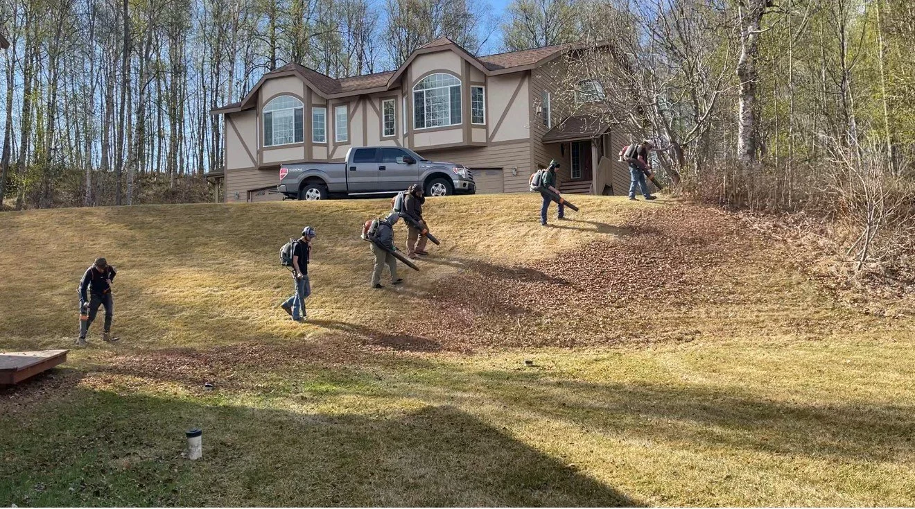 Modern wooden building with large glass window, set against mountainous landscape with dry grass and small trees in foreground.