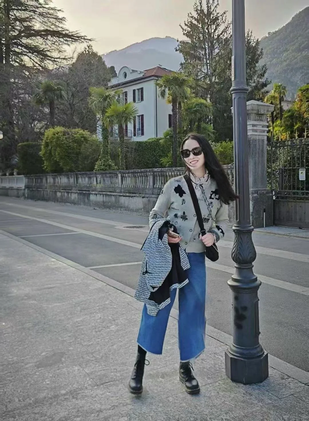 A young woman with long dark hair wearing sunglasses, a light sweater with black patches, blue wide-leg jeans, and black boots, standing on a sidewalk next to a streetlamp. She is holding a jacket and a phone, and smiling at the camera.