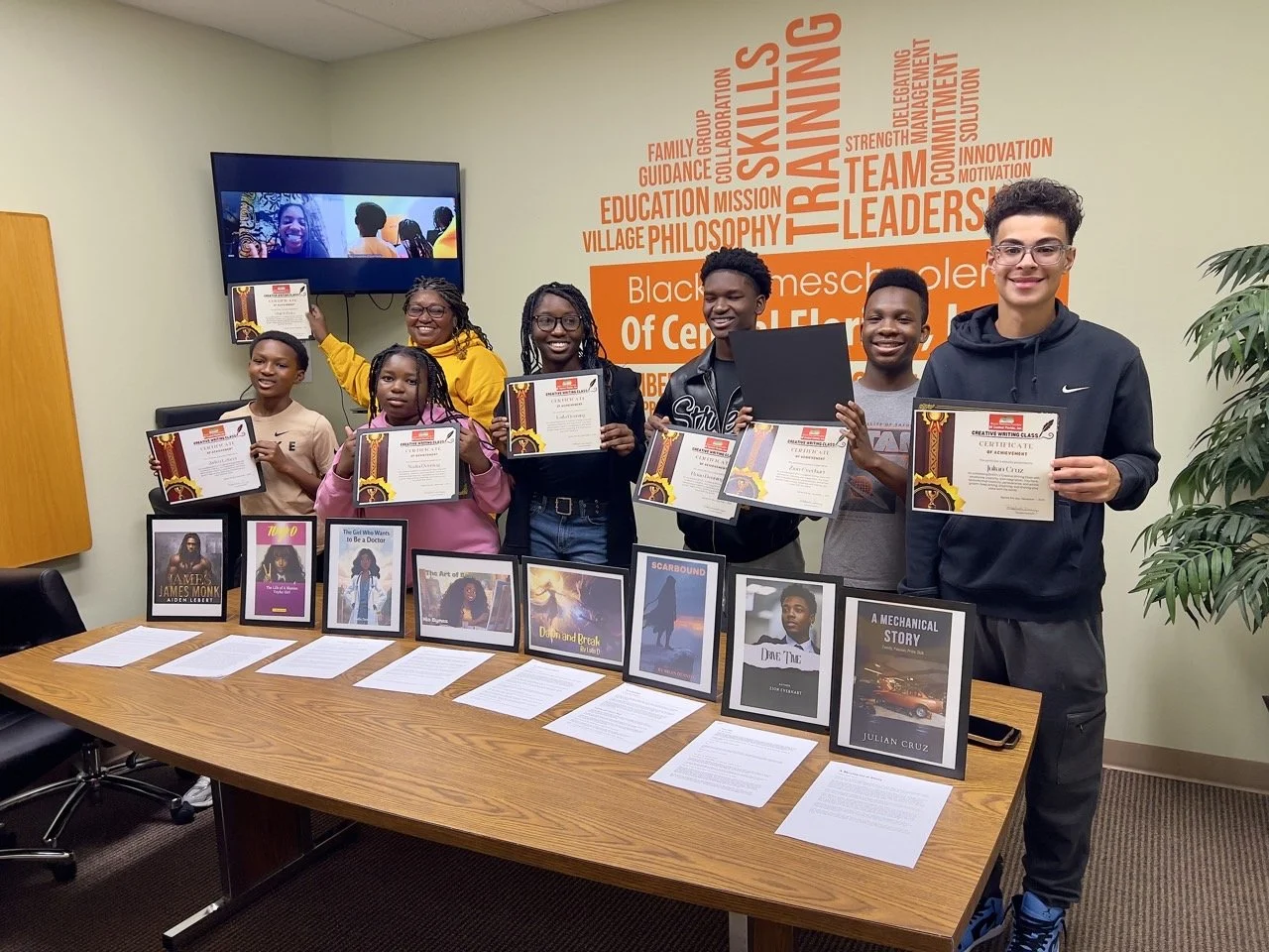 Group of young students holding certificates at a recognition event in a classroom with a table displaying framed book covers and documents. Background features a wall with motivational words related to training and leadership.