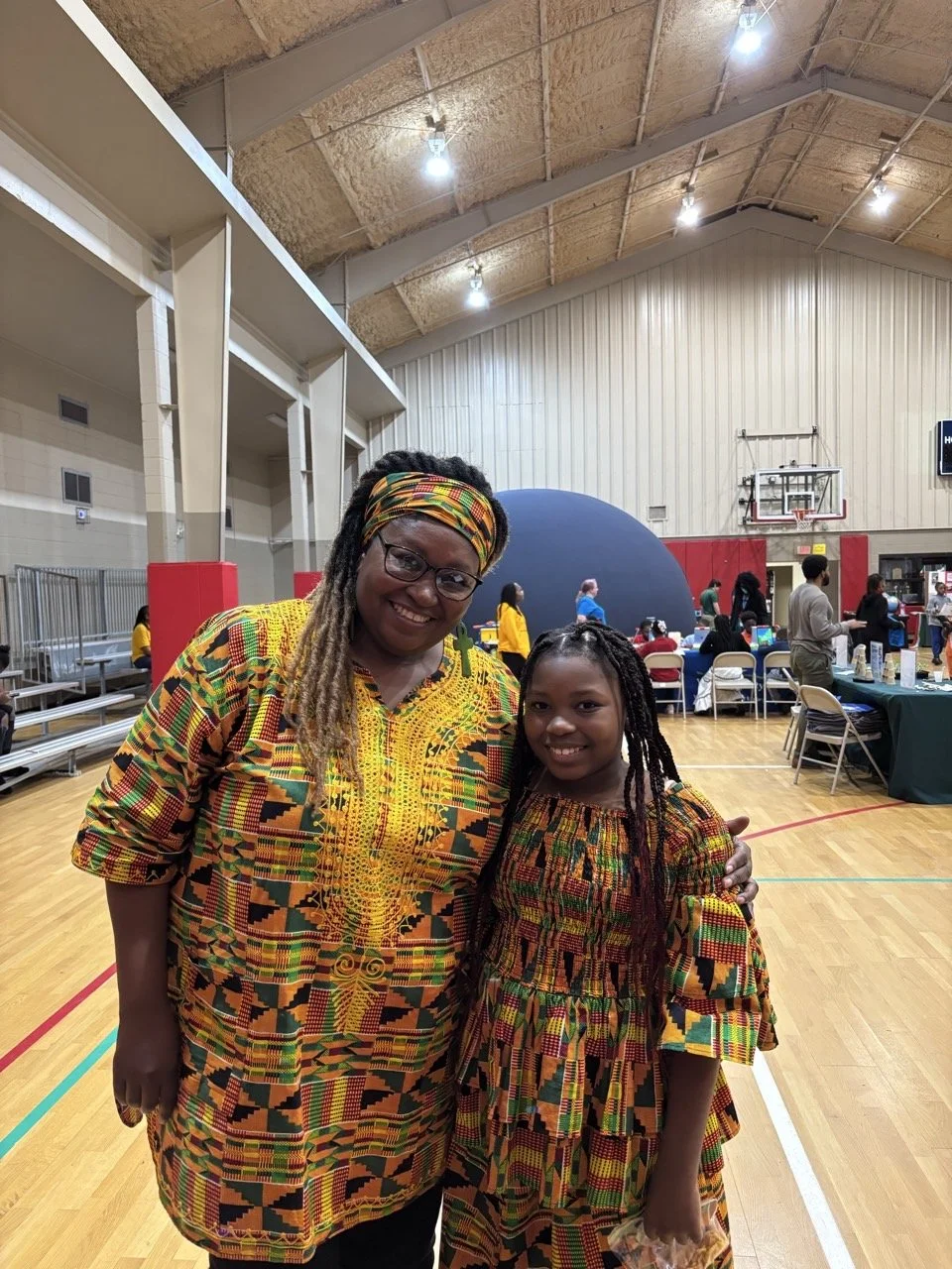 A woman and a girl standing together in a gymnasium, both wearing colorful African-style patterns. The woman has long dreadlocks, glasses, and a headwrap, smiling at the camera. The girl has long braids, smiling, and holding a small item in her hand. In the background, there are tables with people, a large inflatable sphere, and a basketball hoop, with bright indoor lighting.