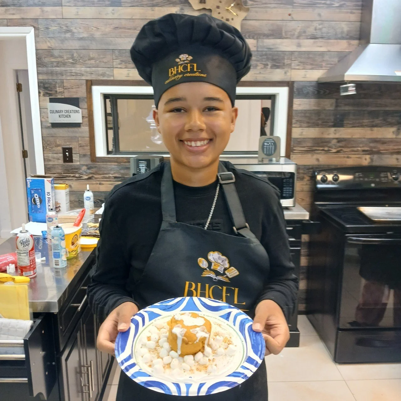 A young person smiling and wearing a chef's hat and apron, holding a plate with dessert topped with sauce and garnishes in a kitchen setting.