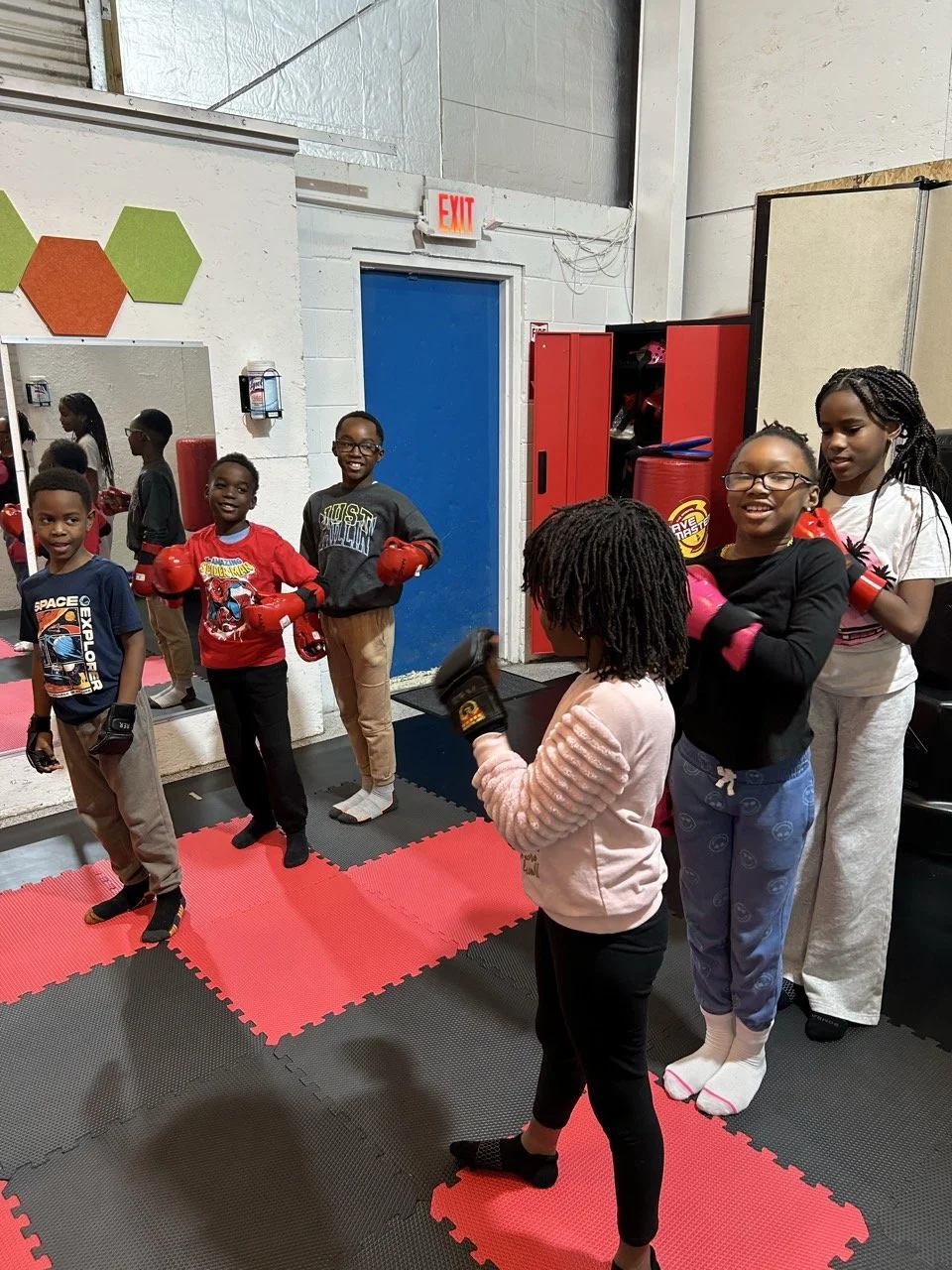 Children in a karate class wearing boxing gloves, practicing in a gym with padded flooring, mirrors, and red cabinets.