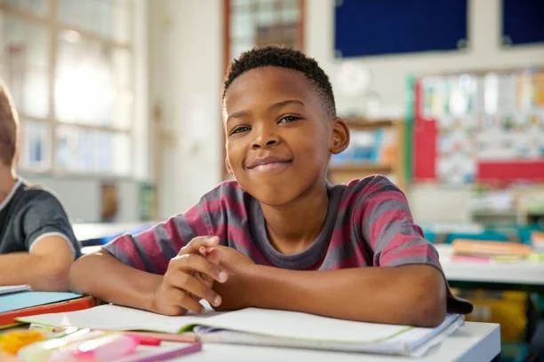 Young boy smiling at school desk with books and stationery.