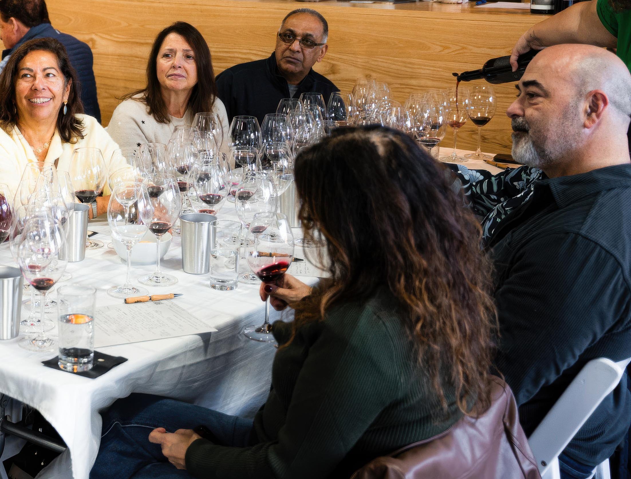 People sitting at a table with numerous wine glasses, some filled with red wine, during a wine tasting event.