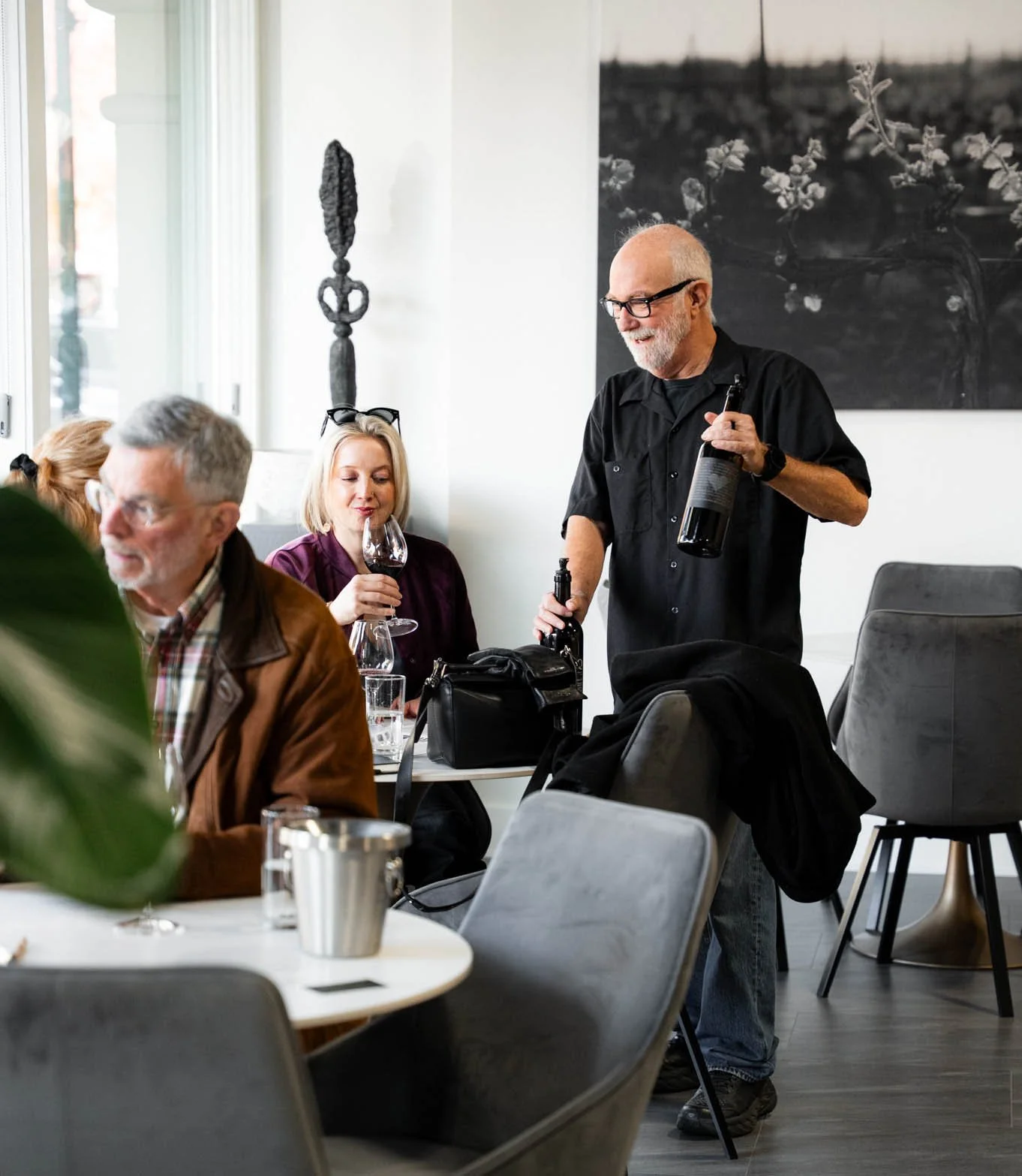 A group of people dining in a restaurant, with a man standing and pouring wine while others, a woman and an older man, sit at the table holding glasses of wine. The restaurant has modern decor with black and white artwork and African tribal art on the wall.