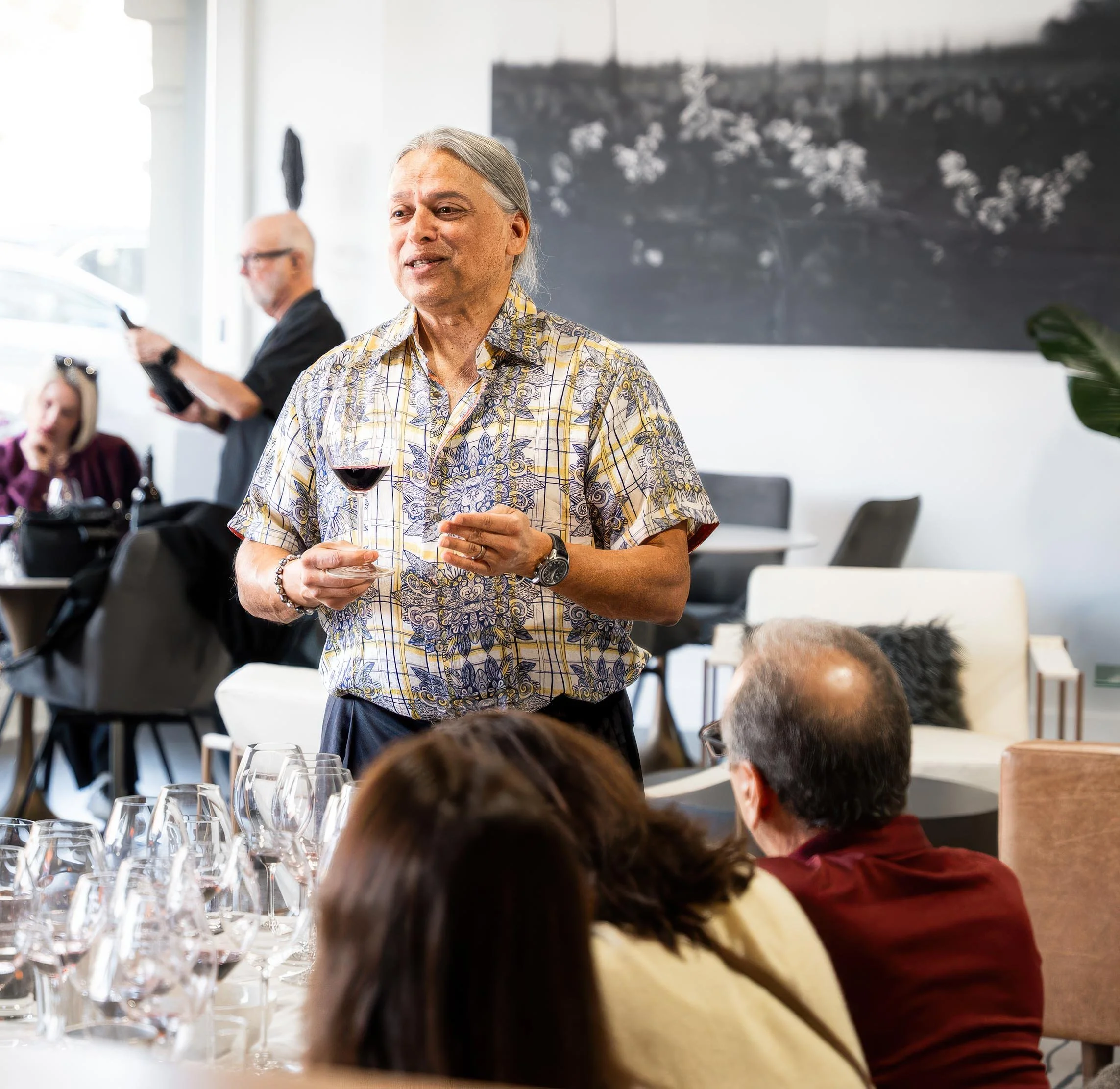 A man with gray hair wearing a patterned short sleeve shirt holding a glass of red wine during a social gathering or meeting in a bright, modern space.