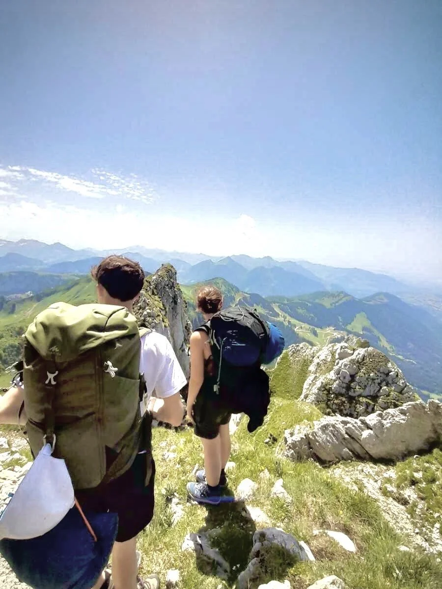 Deux randonneurs sur un sentier de montagne avec des sacs à dos, vue sur les sommets environnants et un ciel dégagé.