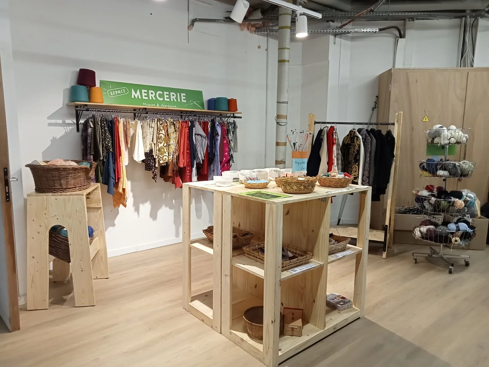 Intérieur d'un magasin de mercerie avec des tissus colorés suspendus, un présentoir avec des paniers, des étagères en bois avec des articles de couture, et un stand de pelotes de laine. "Espace Mercerie" est affiché sur un panneau vert.