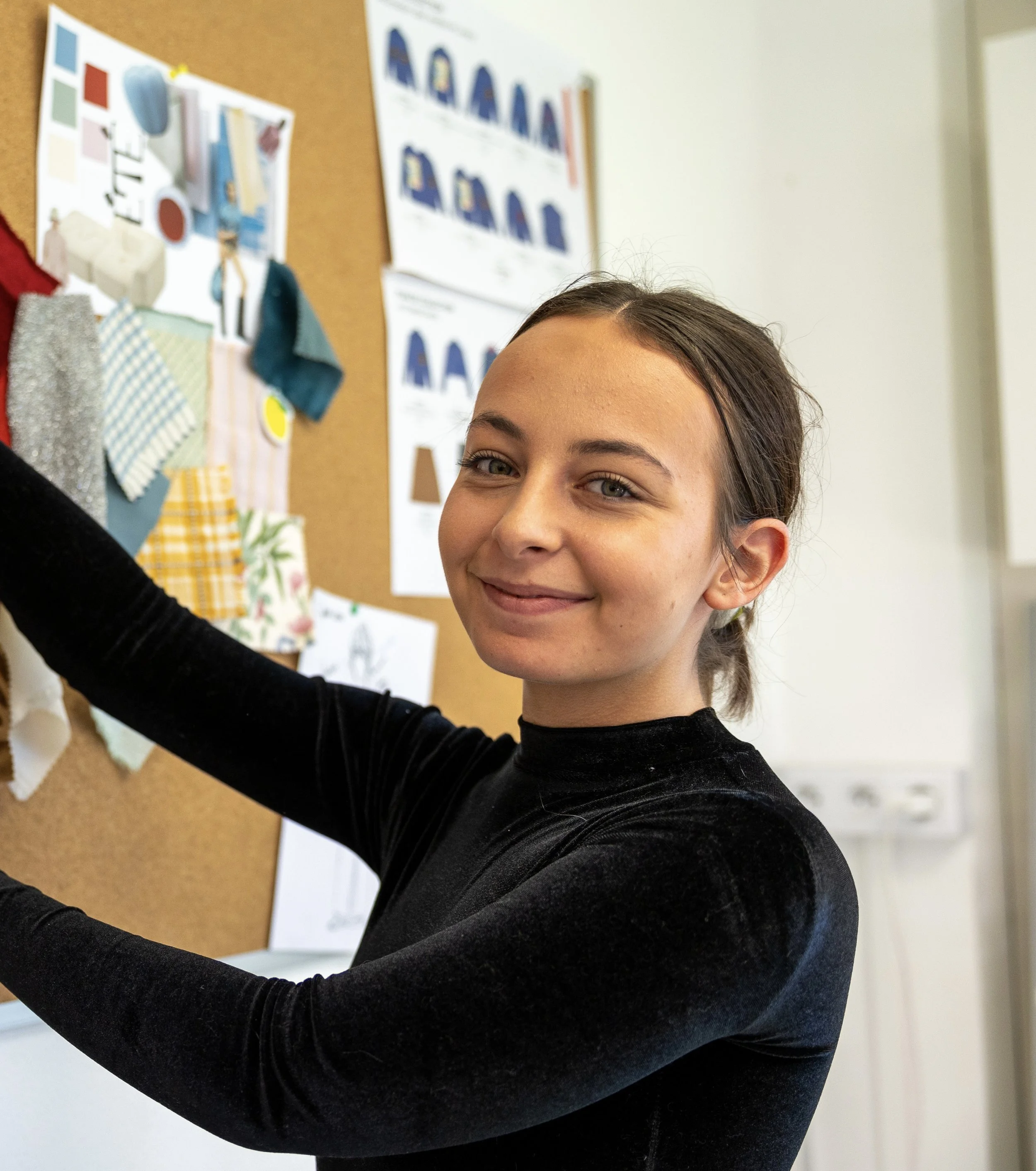 Une femme souriante épinglant des échantillons de tissus sur un tableau d'affichage, avec des croquis et des motifs en fond.