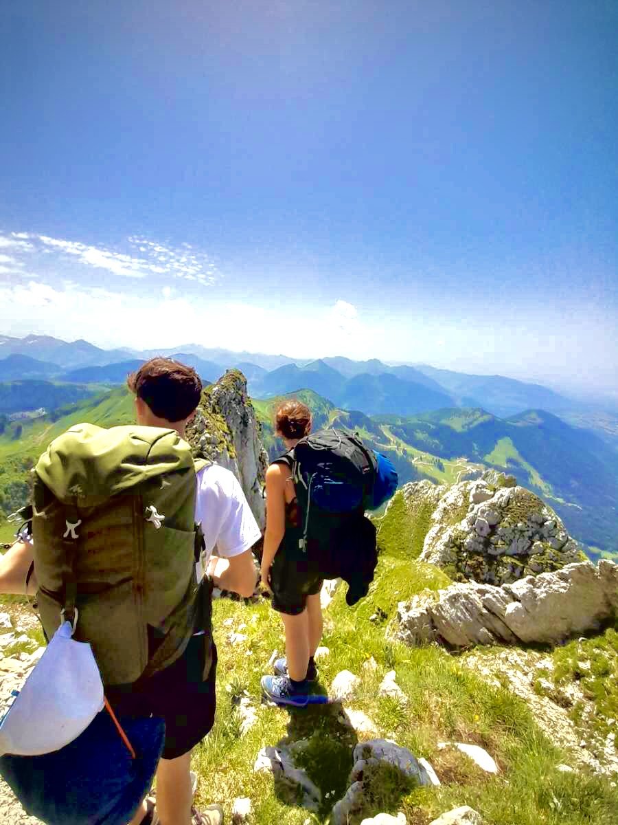 Deux personnes avec des sacs à dos marchent sur un sentier de montagne, avec une vue panoramique sur les sommets et le ciel bleu.